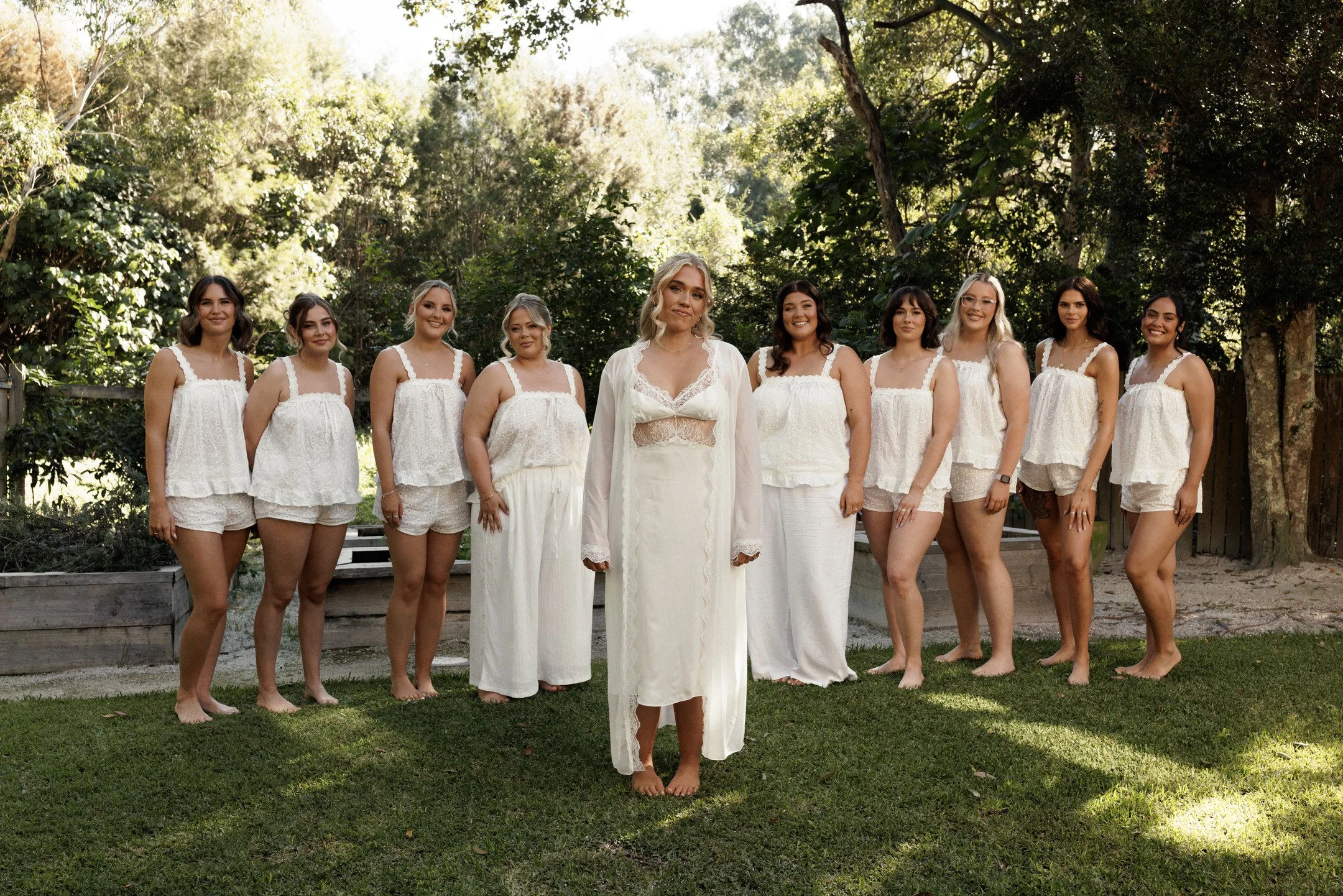 A group of women, dressed in white, standing outdoors in front of trees.