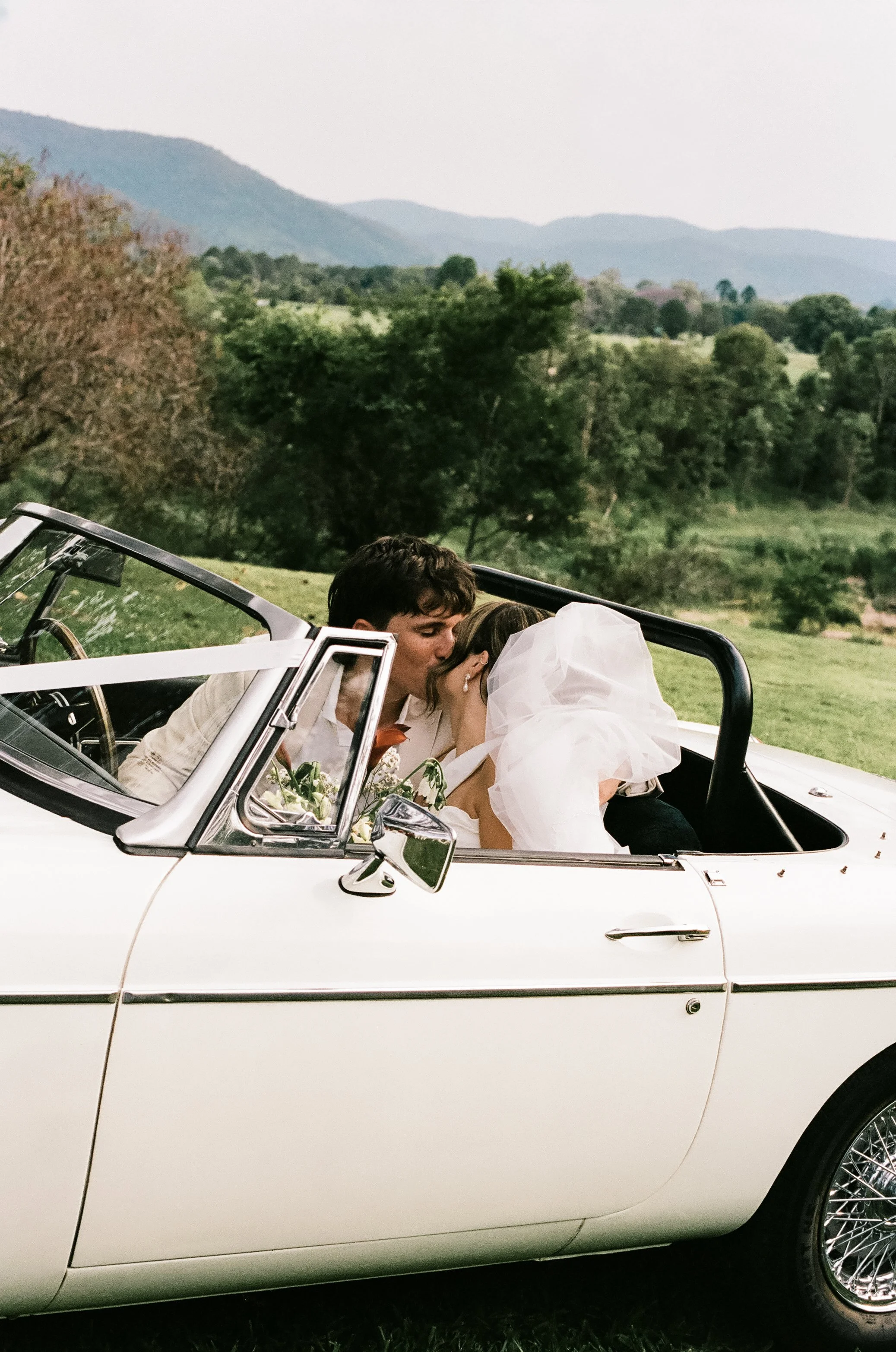 A bride and groom sharing a kiss while sitting in a vintage white convertible car outdoors, with lush green trees and mountains in the background.