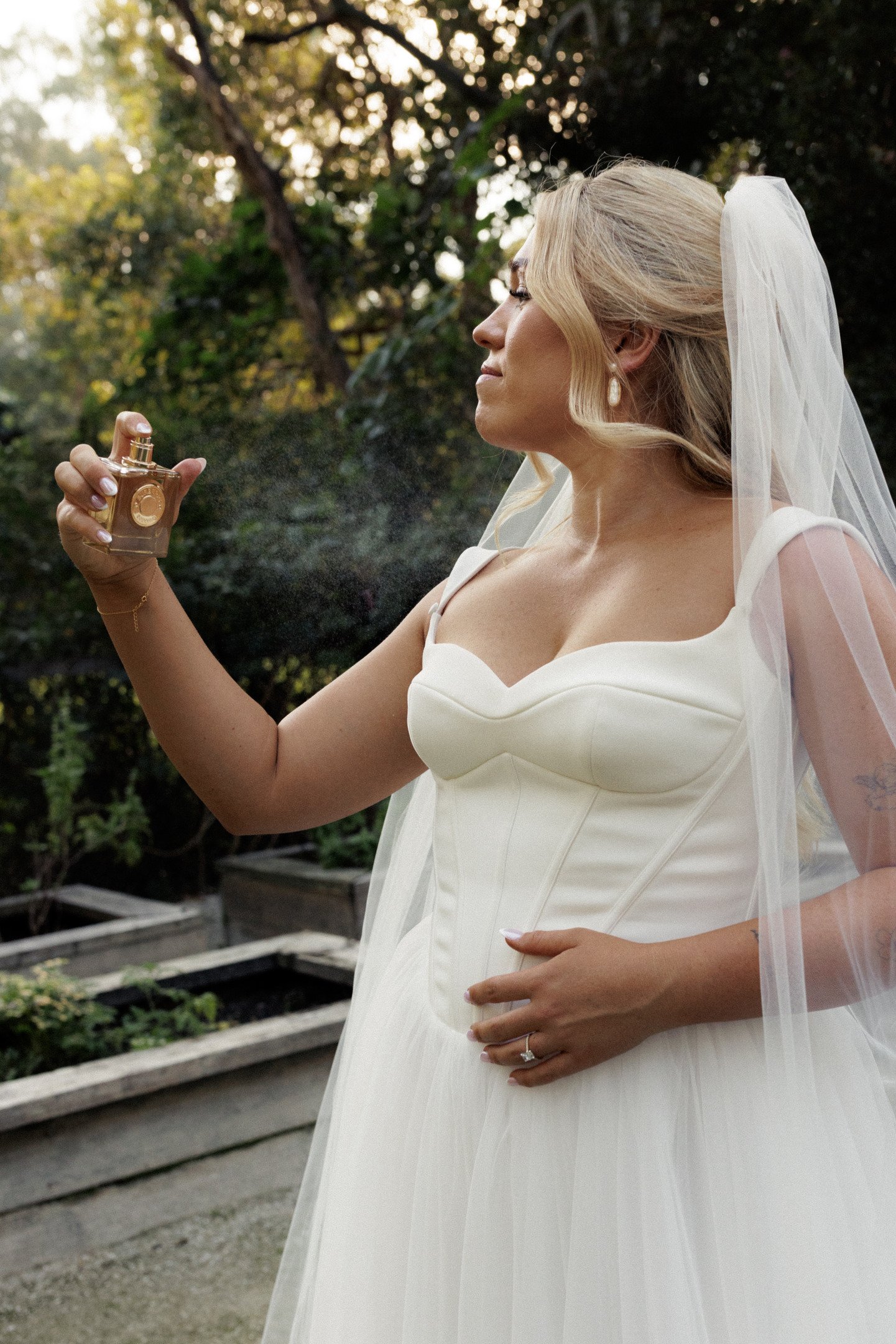 A bride in a white wedding dress and veil holding a perfume bottle outdoors during daytime.