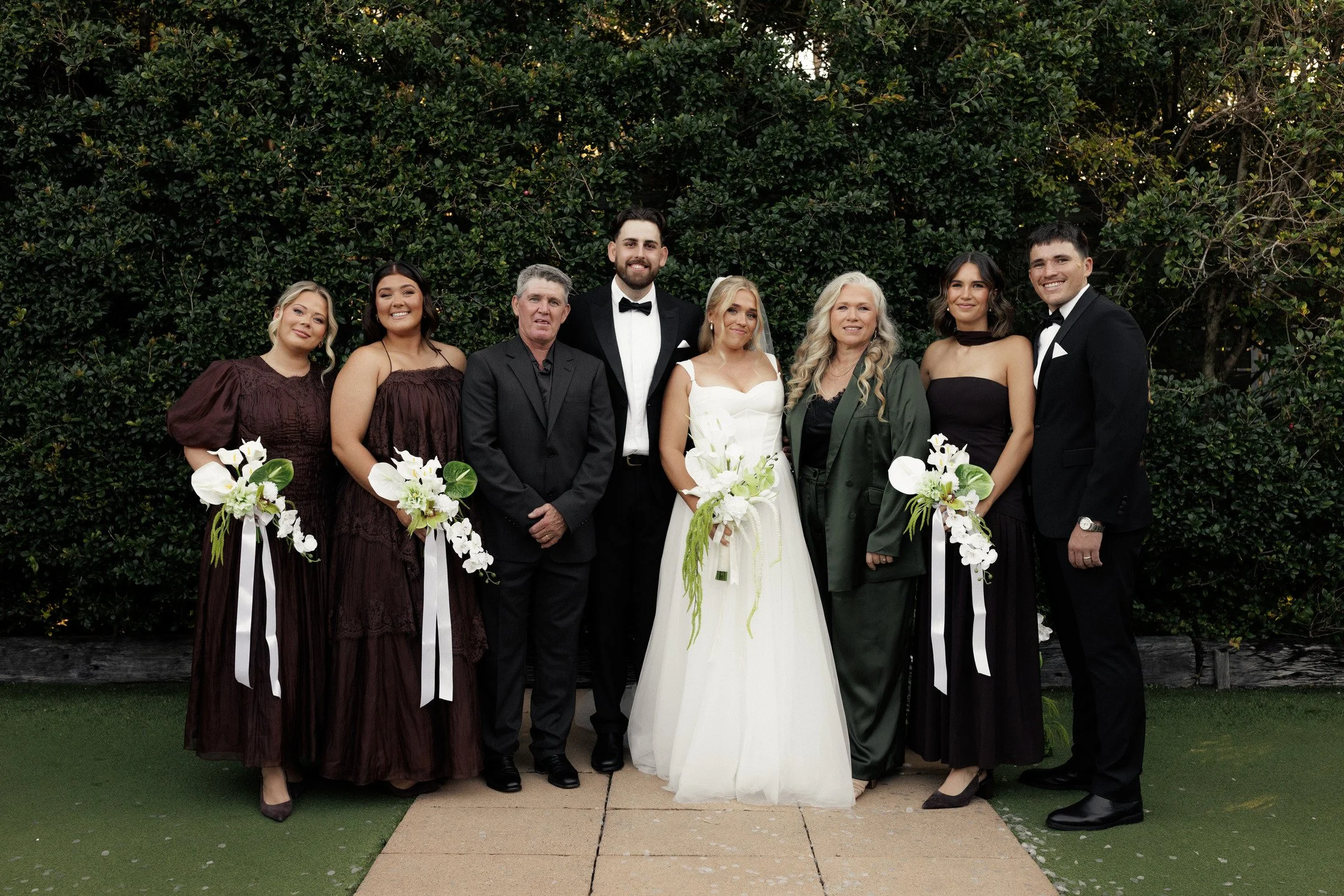 Group of eight people, including a bride and groom, posing outdoors in front of greenery for a wedding photo. The bride is in a white wedding dress holding a bouquet, and the groom is in a black tuxedo with a bow tie. Other guests are dressed in eleg