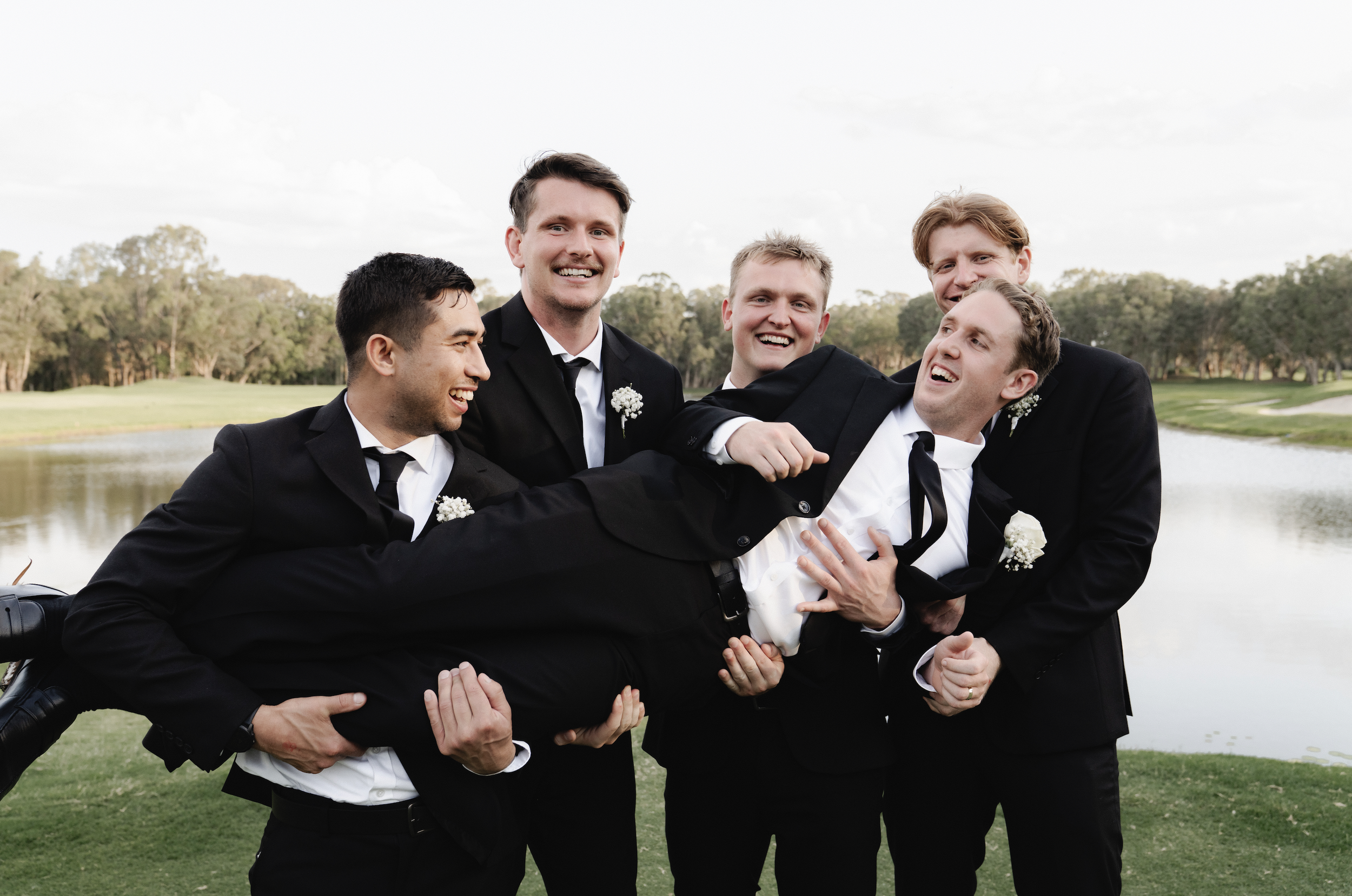 Group of groomsmen in black suits playing around outdoors, one being held up by the others, near a pond with trees in the background - Elevated Weddings shot on 35mm film