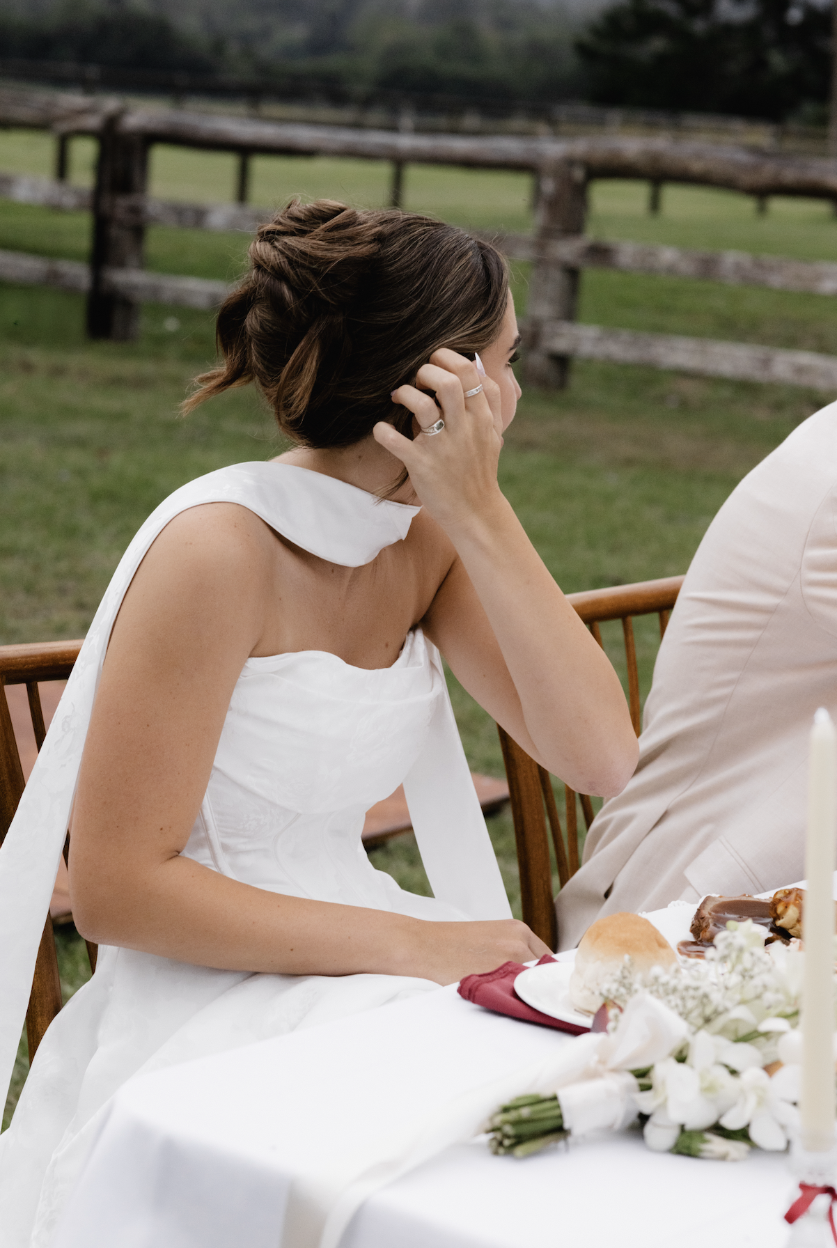 A woman in a white wedding dress sitting at a table with a bouquet of white flowers, a plate of food, and a lit candle, outdoors near a wooden fence.