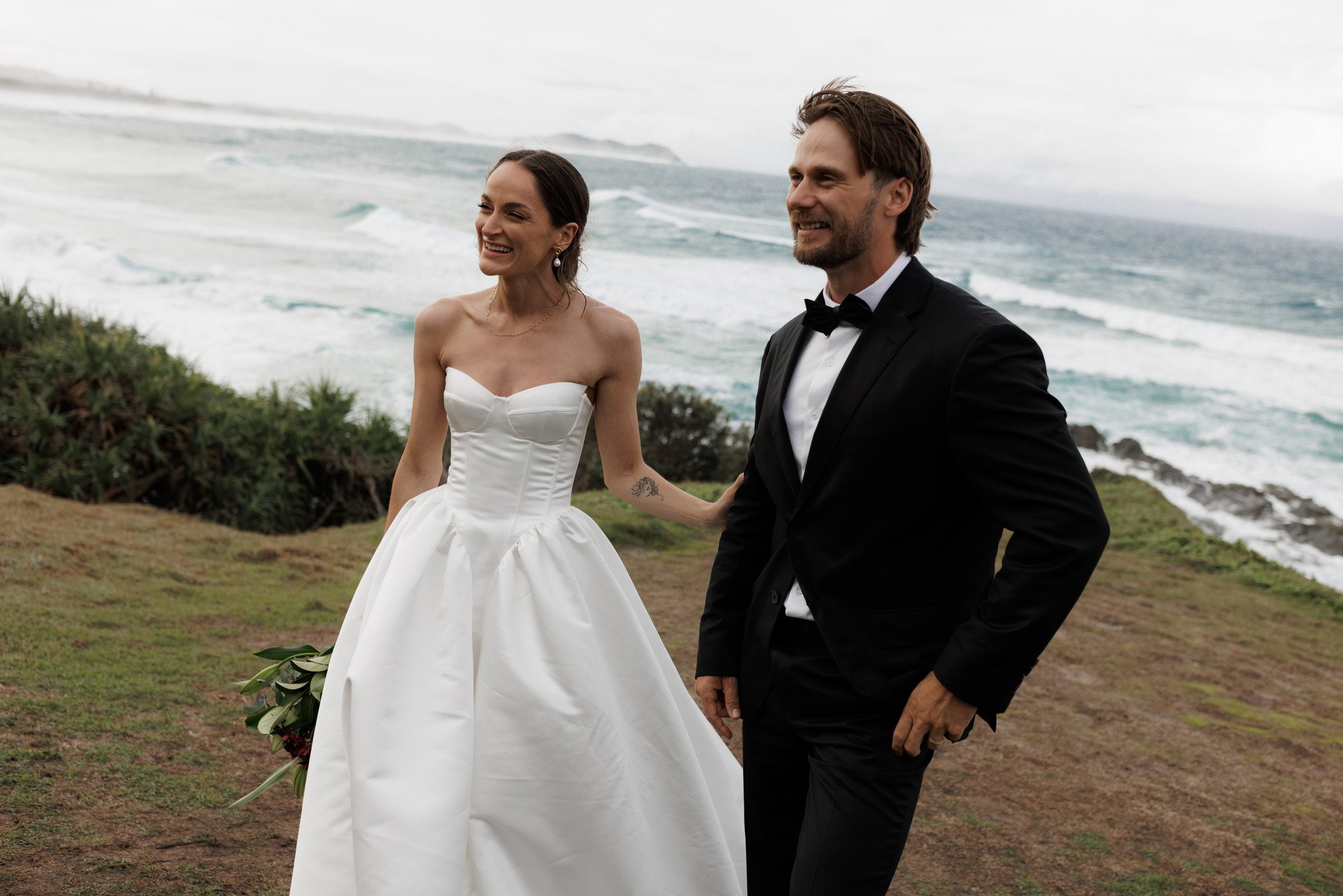 A couple dressed in wedding attire, smiling, holds hands on a grassy area near the ocean with waves and overcast sky in the background.