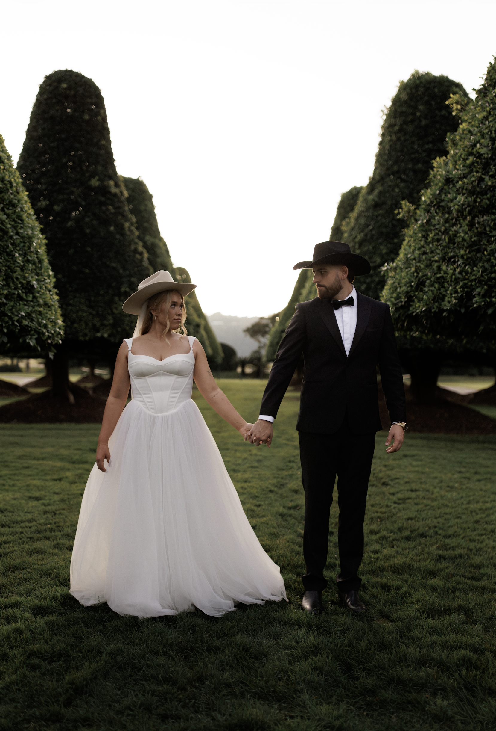 A bride and groom holding hands walking on a grassy field with tall, well-trimmed trees in the background during sunset.