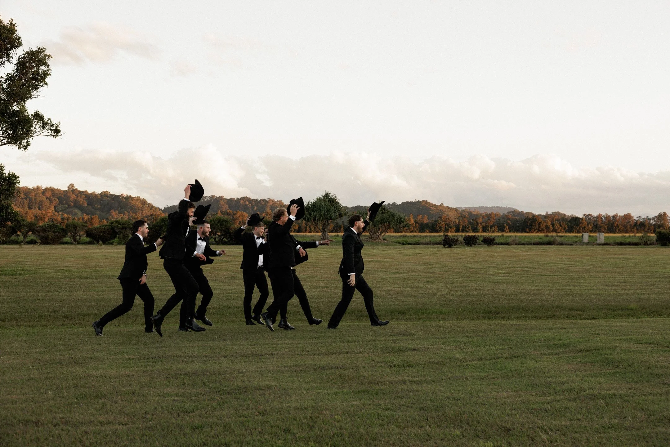A group of men in formal black suits and tuxedos, with some lifting their black hats, walking across a grassy field during late afternoon or early evening. In the background, there are trees, hills, and a partly cloudy sky.