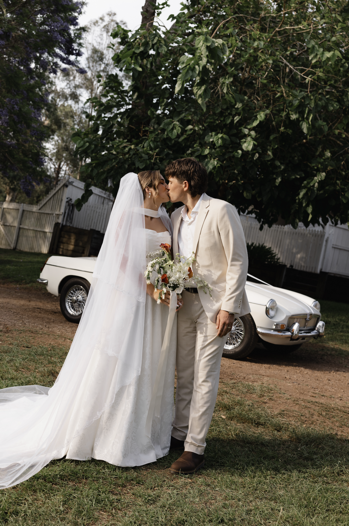 A bride and groom kiss outdoors, with a vintage white car and lush green foliage in the background.