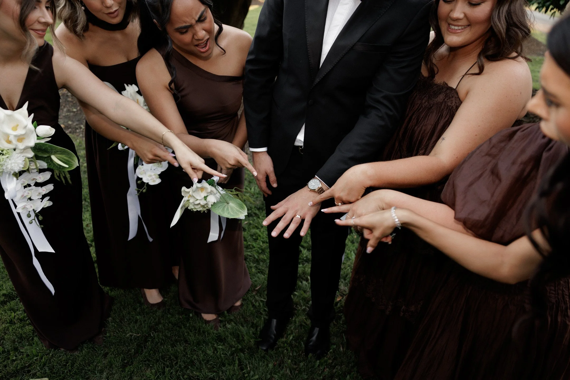 A group of women and a man at a wedding ceremony, raising and pointing their fingers towards a wedding cake.