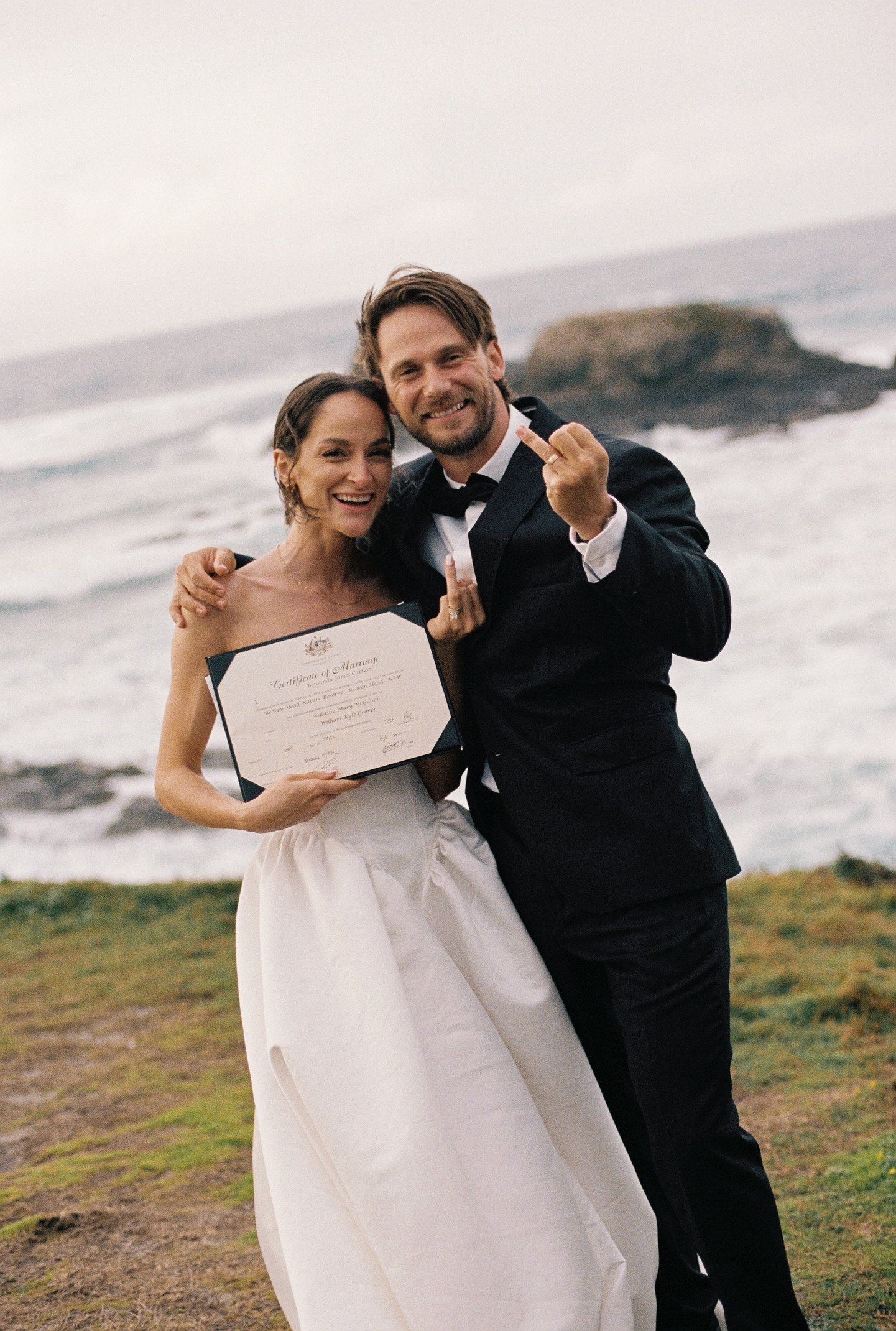 A couple in wedding attire celebrating on a beach, holding a marriage certificate and flashing middle fingers.