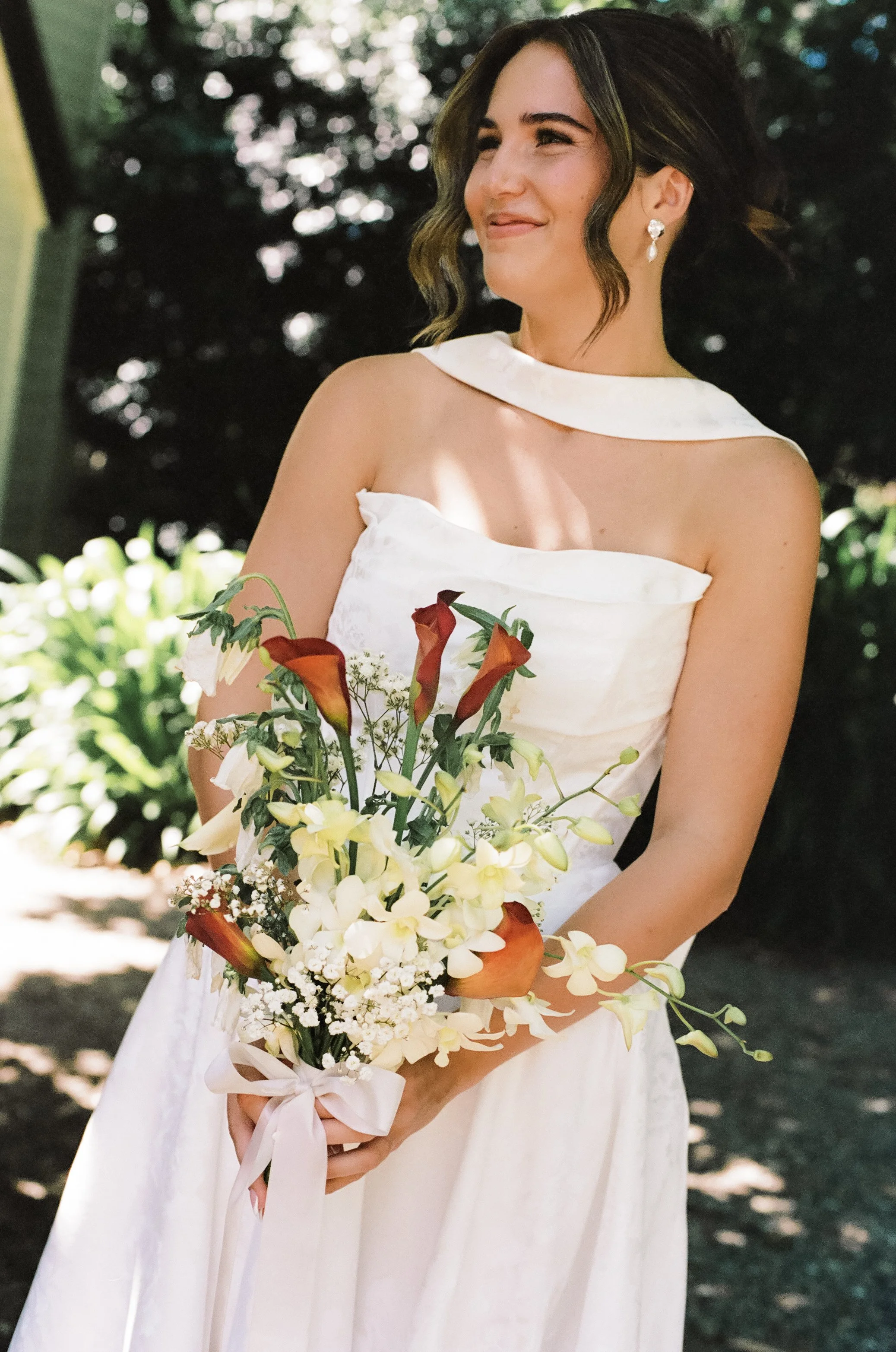 A woman in a white wedding dress holding a bouquet of red and white flowers outdoors.