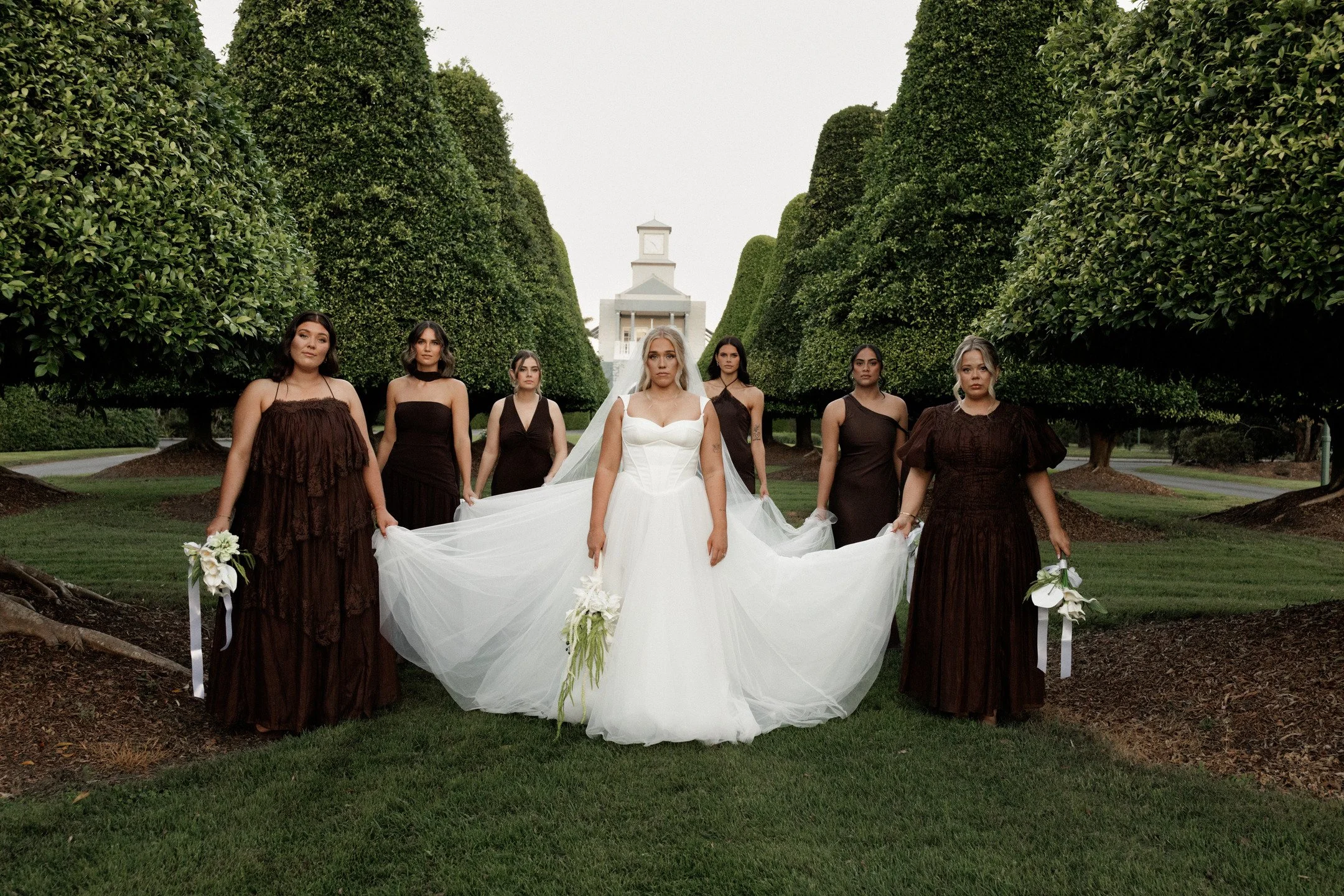 A bride in a white wedding dress and six bridesmaids in dark brown dresses standing outdoors among tall green bushes, with a white building and clock tower in the background.