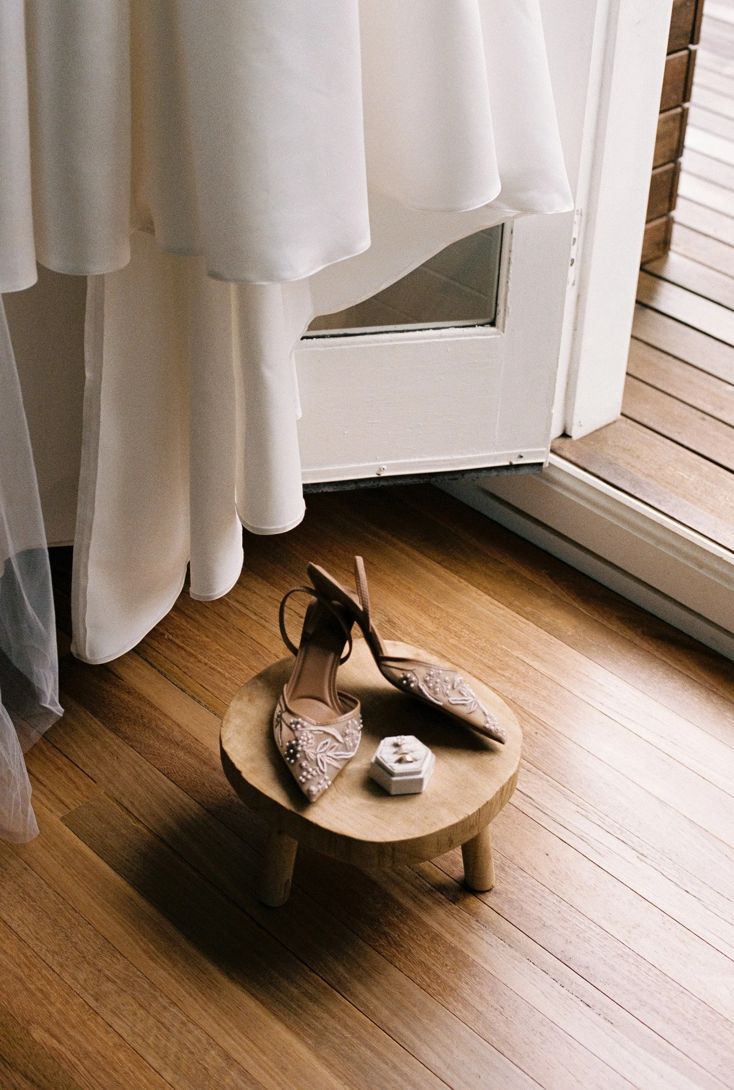 A pair of embellished high-heeled shoes on a small wooden stool, with a white curtain and a door with a glass panel in the background.