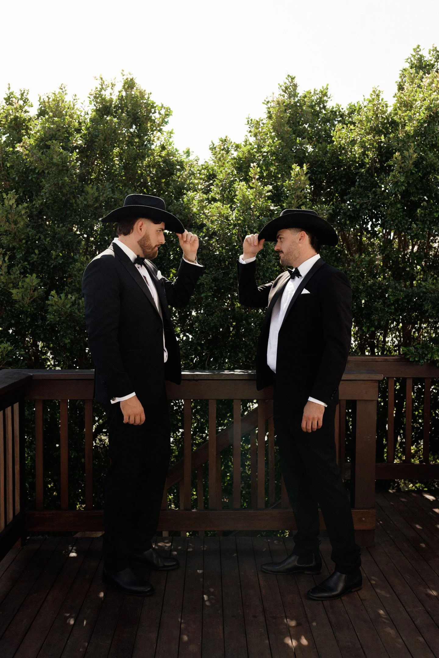 Two men dressed in black tuxedos, white shirts, black bow ties, and black cowboy hats, standing on a wooden deck with a railing, facing each other and touching their hats, outdoors with green trees in the background.
