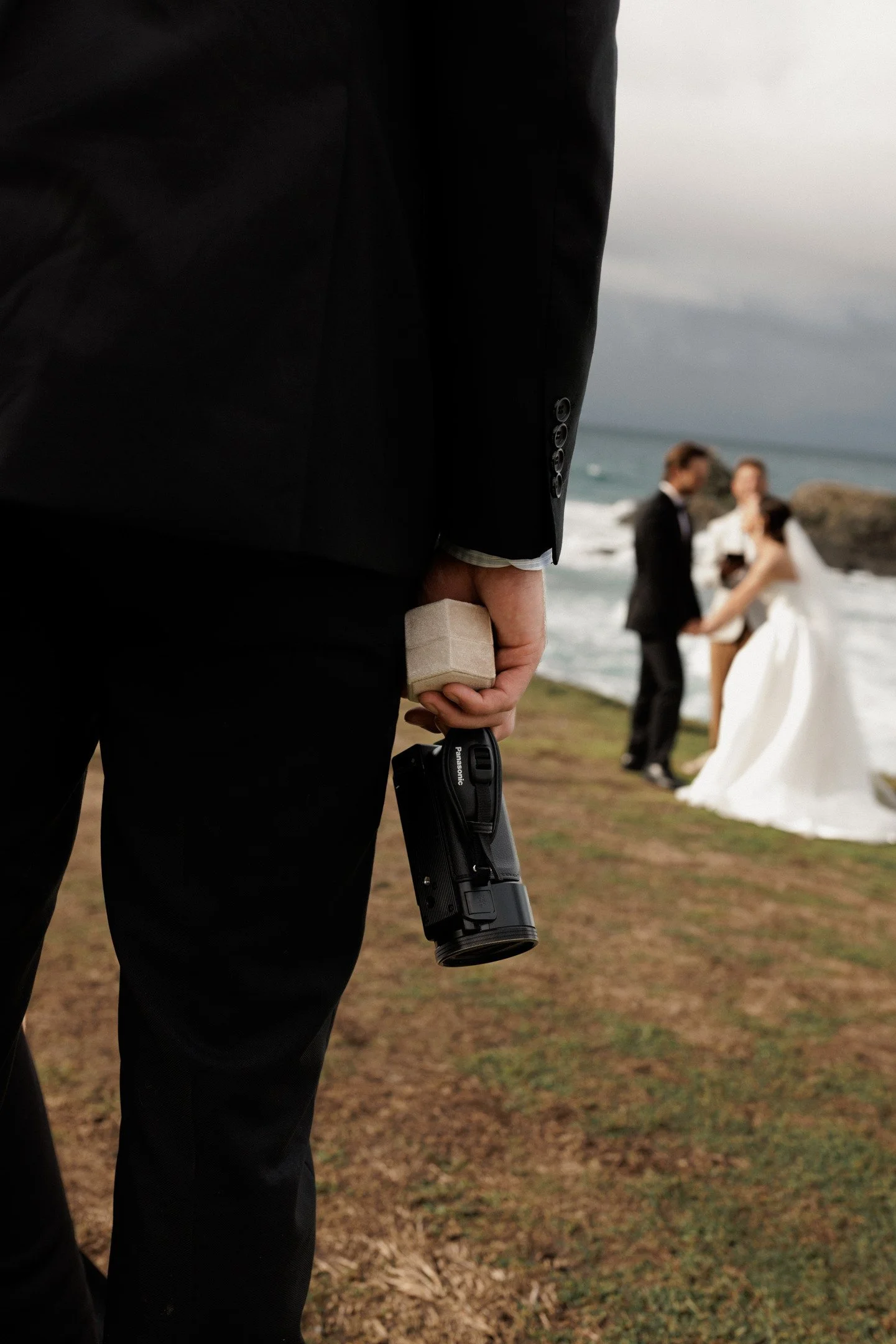 Close-up of a person wearing a black suit holding a small box and a handgun at a beach with a blurred background of a couple in wedding attire.