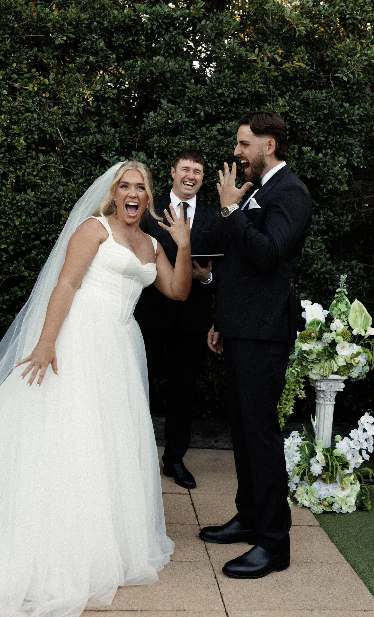 A bride and groom are exchanging vows at a wedding ceremony, with a officiant standing behind them, all laughing and smiling.