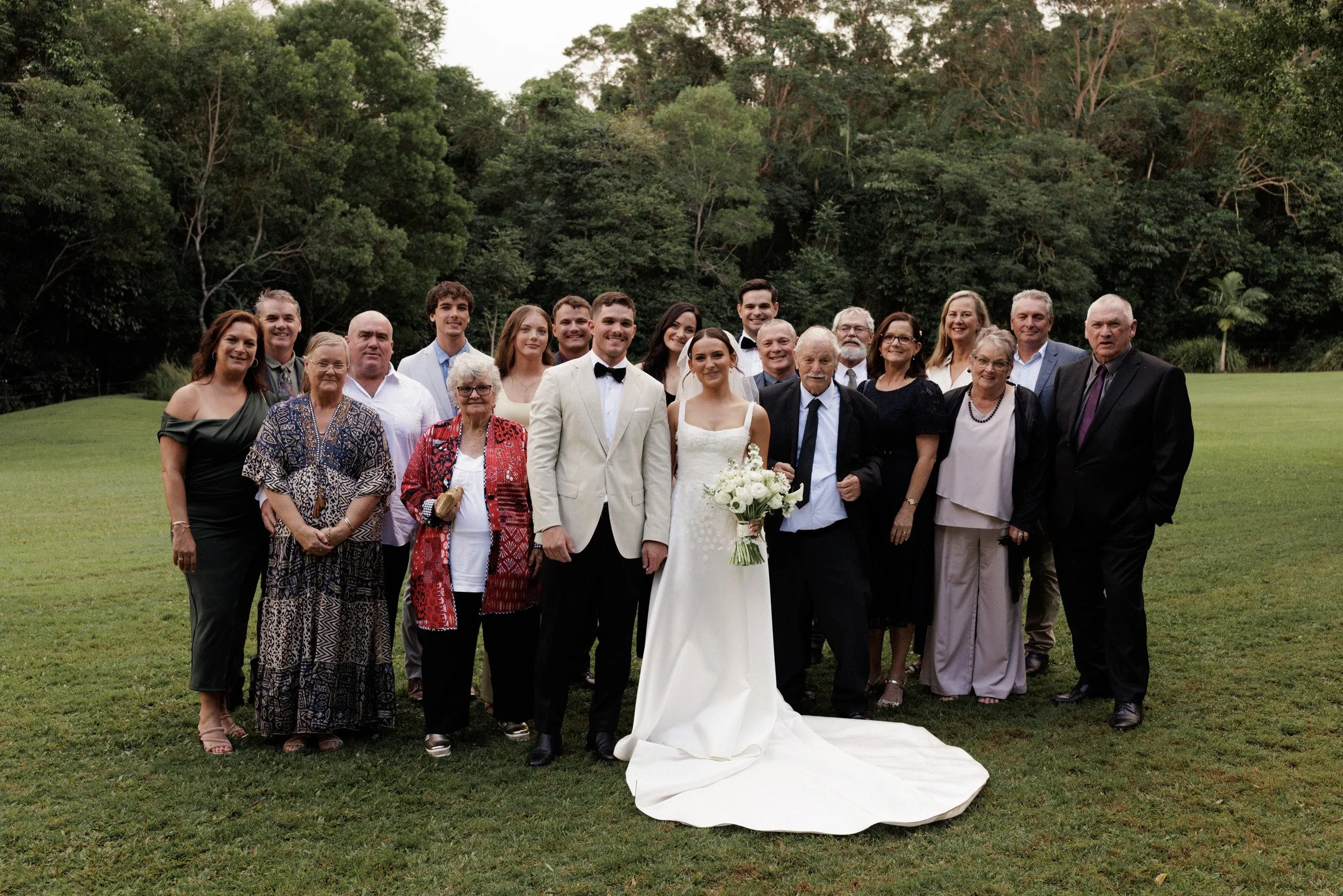 A large group of people, including a bride and groom, posing outdoors on a grassy area with trees in the background. The bride is holding a bouquet of white flowers, and the groom is wearing a light suit with a bow tie.