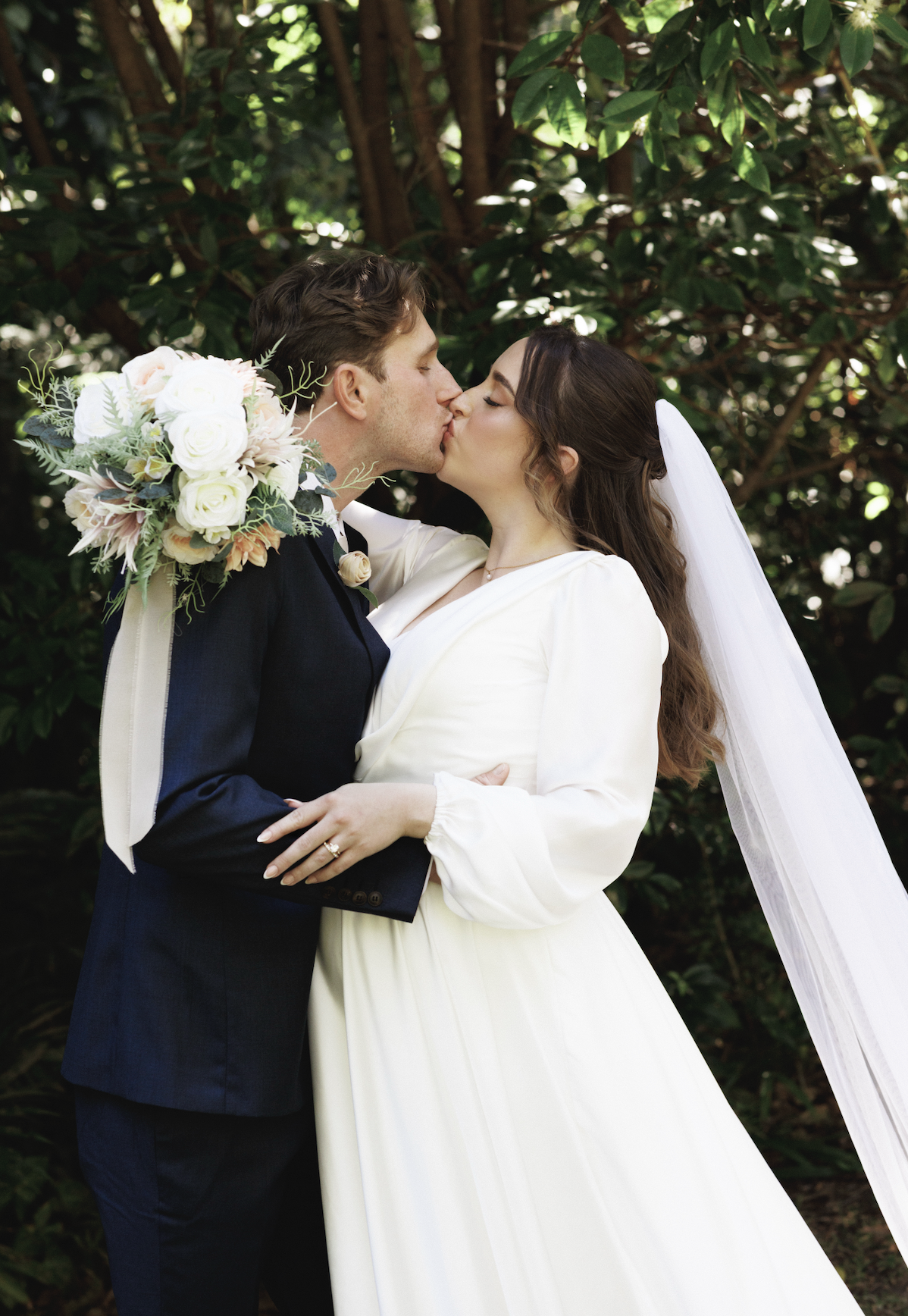 A bride and groom are kissing outdoors, with the groom carrying a bouquet of white and pink flowers over his shoulder. The bride is wearing a white dress and veil, and the groom is in a dark suit. They are surrounded by green foliage.