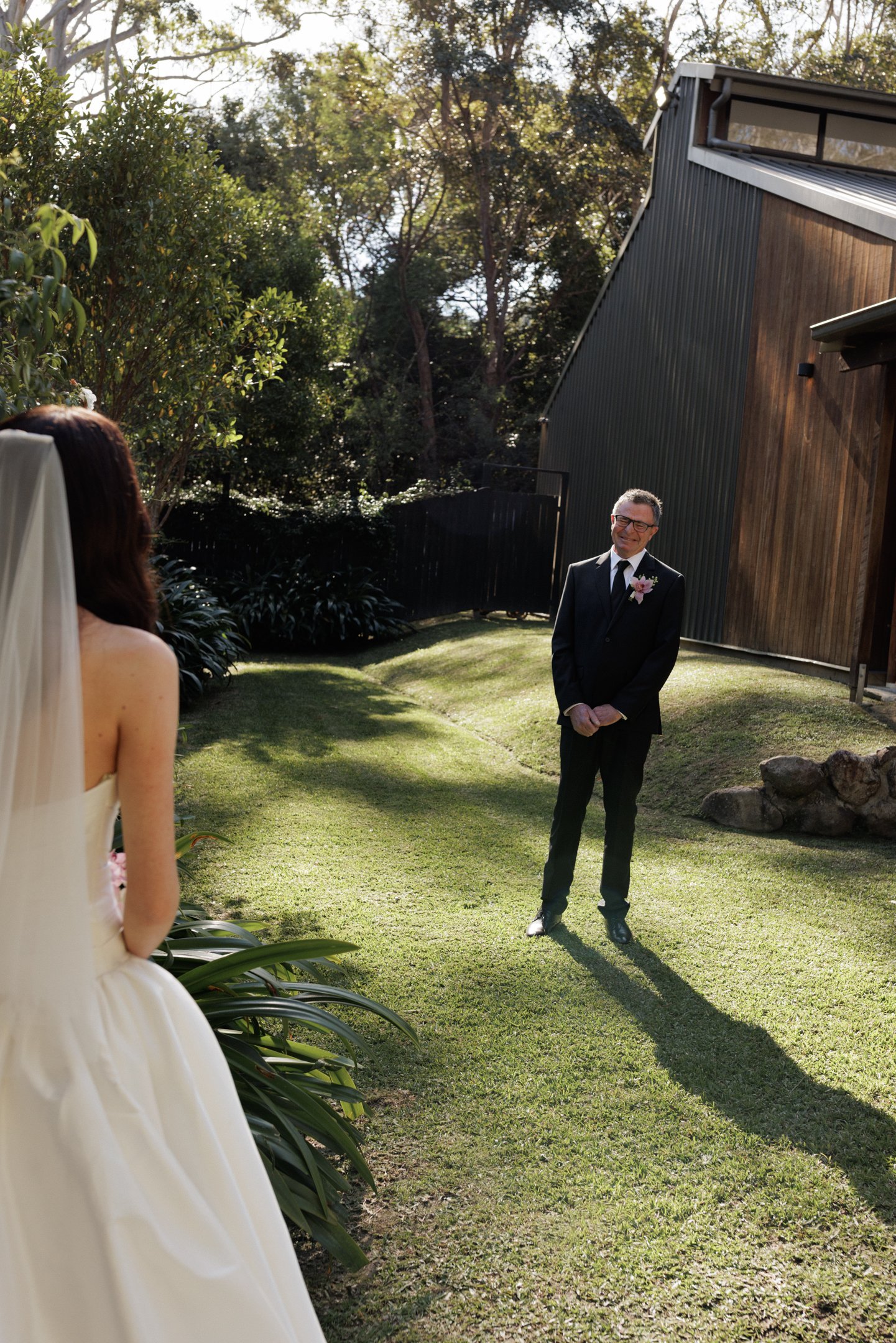 A wedding outdoor ceremony with a woman in a white dress and a man in a black suit, both smiling, on a sunny day in a garden with trees and a wooden building in the background.
