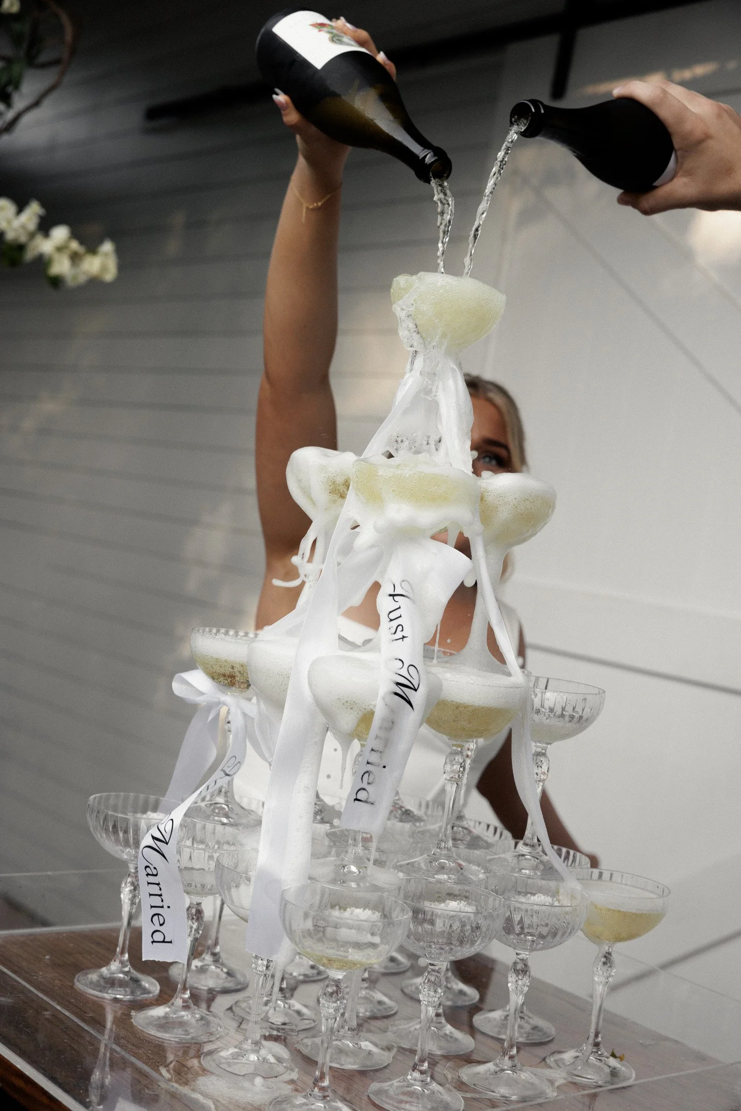 A woman is pouring champagne into a champagne tower made of stacked glasses. The glasses have labels attached to ribbons draped around them.