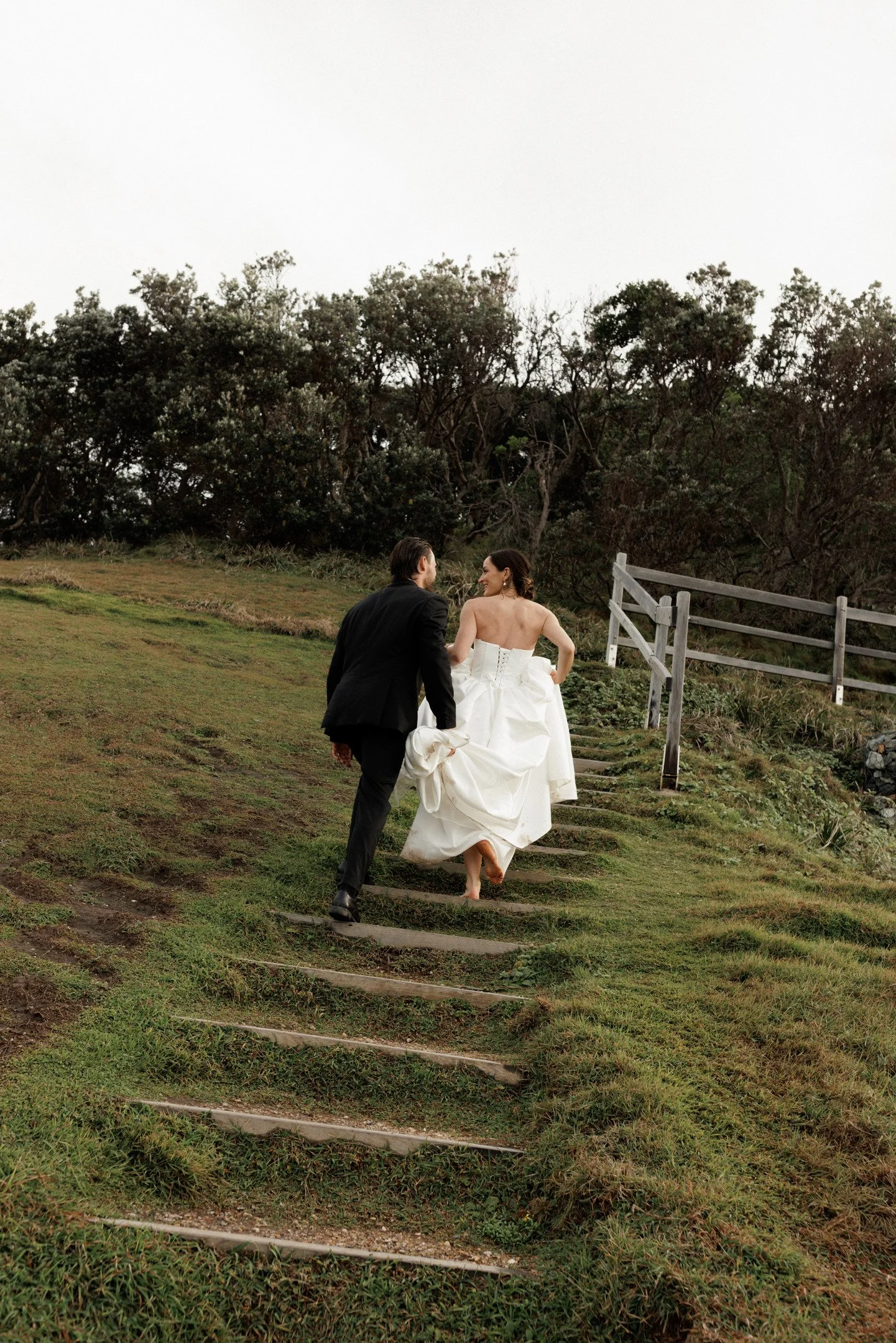 A bride and groom walking up outdoor steps on a grassy hillside with trees in the background. The bride wears a white wedding dress, and the groom wears a black suit.