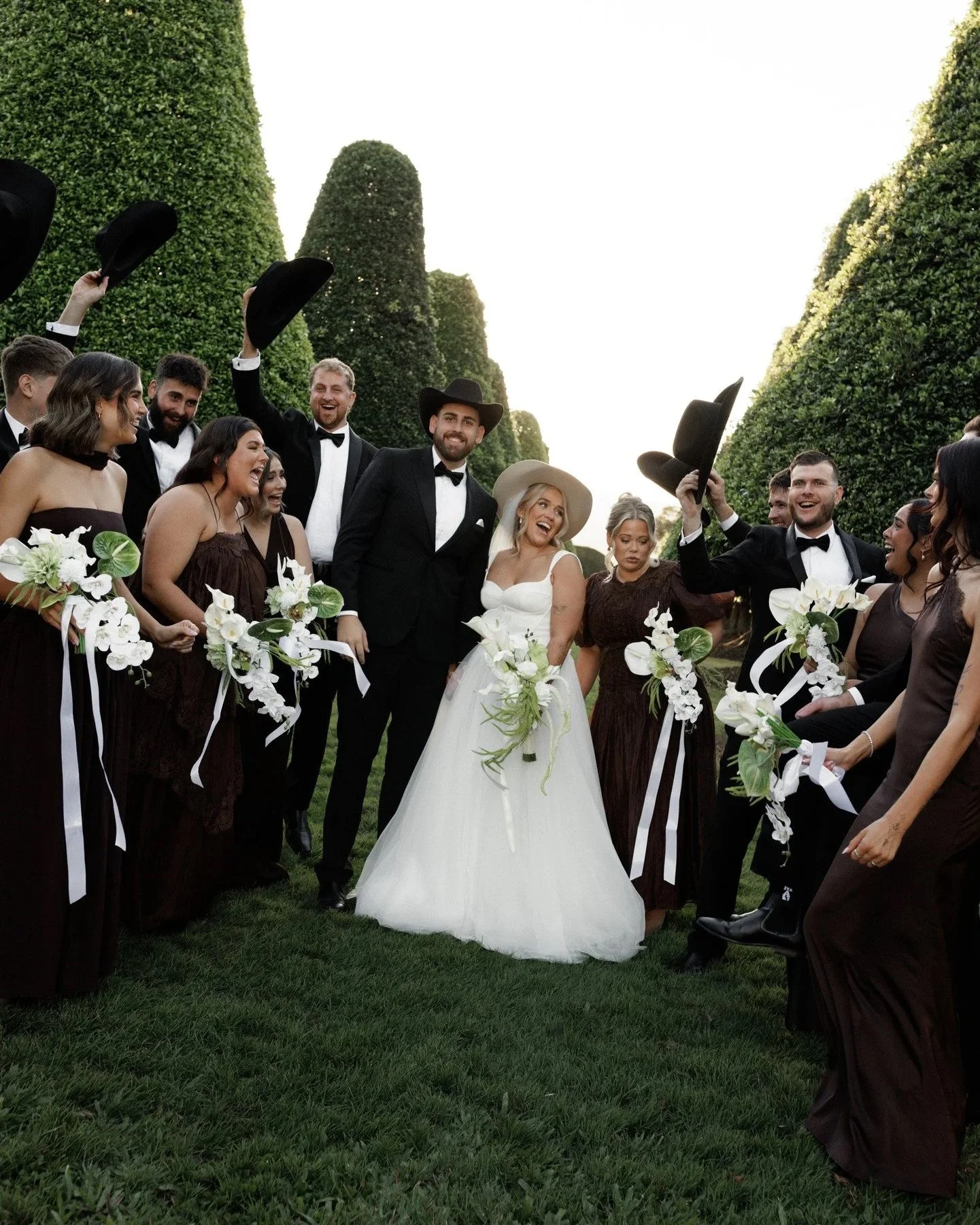 A group of people celebrating a wedding outdoors, with some holding hats in the air, dressed in formal attire, with the bride in a white gown and veil, and the groom in a black tuxedo with a cowboy hat, surrounded by friends and family holding white floral bouquets and standing on green grass under a tall hedge.