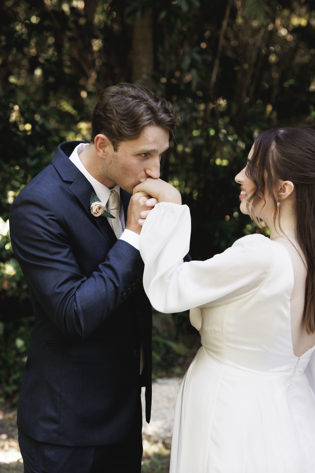 A groom in a navy suit kissing a bride's hand outdoors. The bride wears a white dress with long sleeves, and they are surrounded by green foliage.