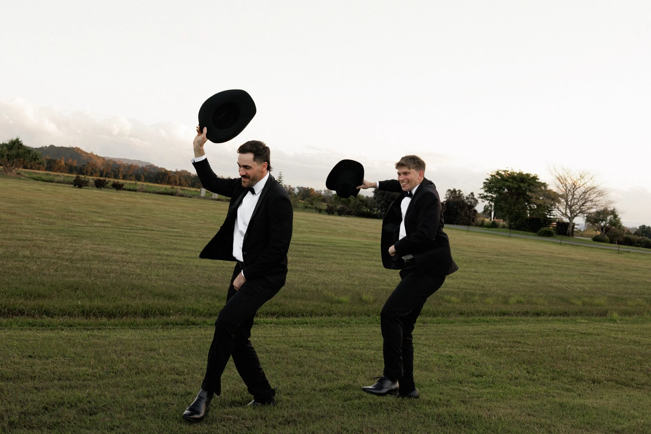 Two men dressed in tuxedos and dress shoes playfully dancing outdoors on a grassy field, holding hats in the air, with trees and hills in the background.