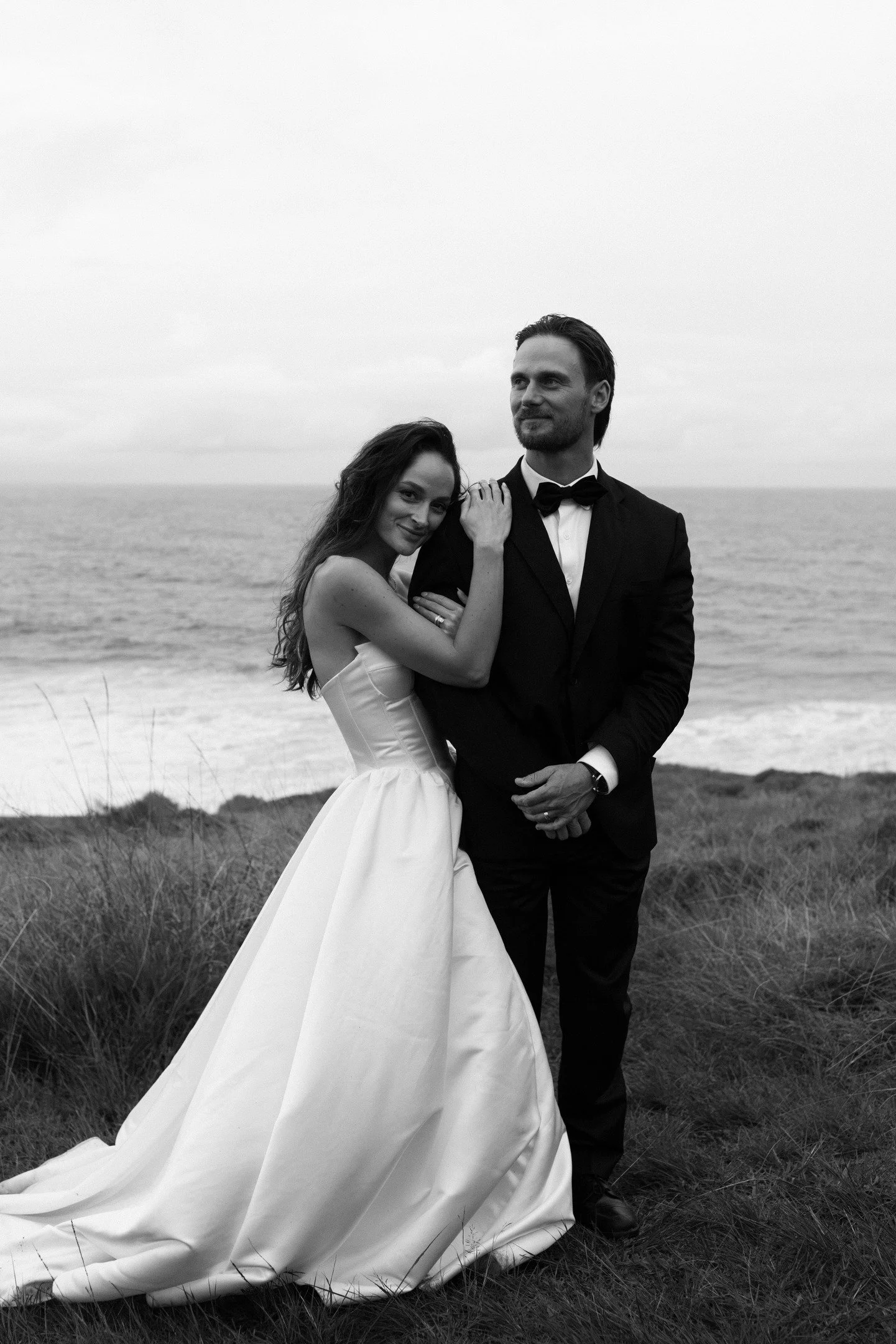 A black-and-white photo of a bride and groom on a beach. The bride is leaning on the groom's shoulder, smiling, and wearing a strapless wedding gown with a long train. The groom is dressed in a tuxedo, standing with his hands clasped in front of him,