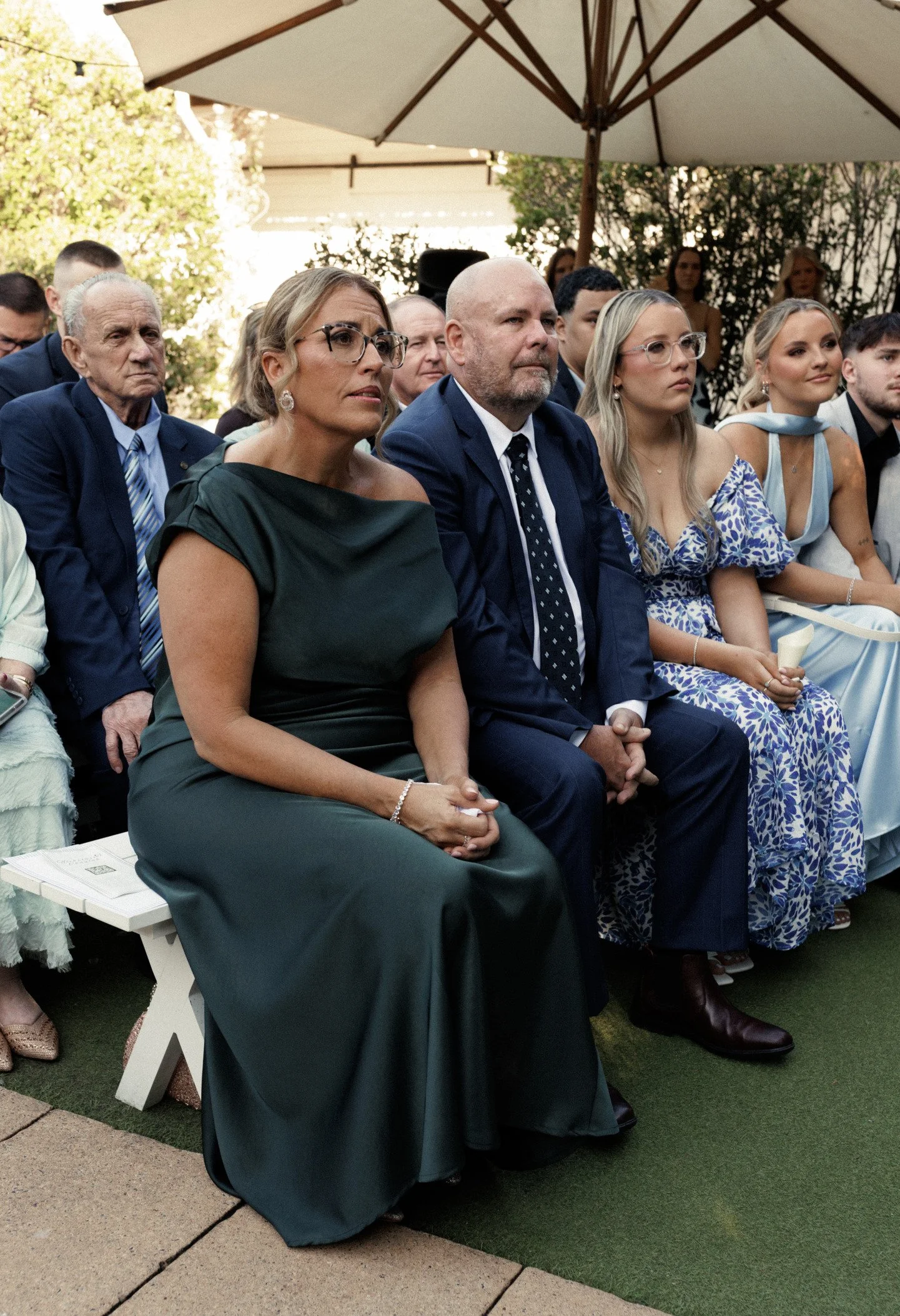 Group of people attending outdoor event, seated in chairs under large umbrellas. Including elderly man, woman in dark dress, man in suit with tie, women in dresses, and other attendees. Some are focused, others smiling. Greenery and trees in backgrou