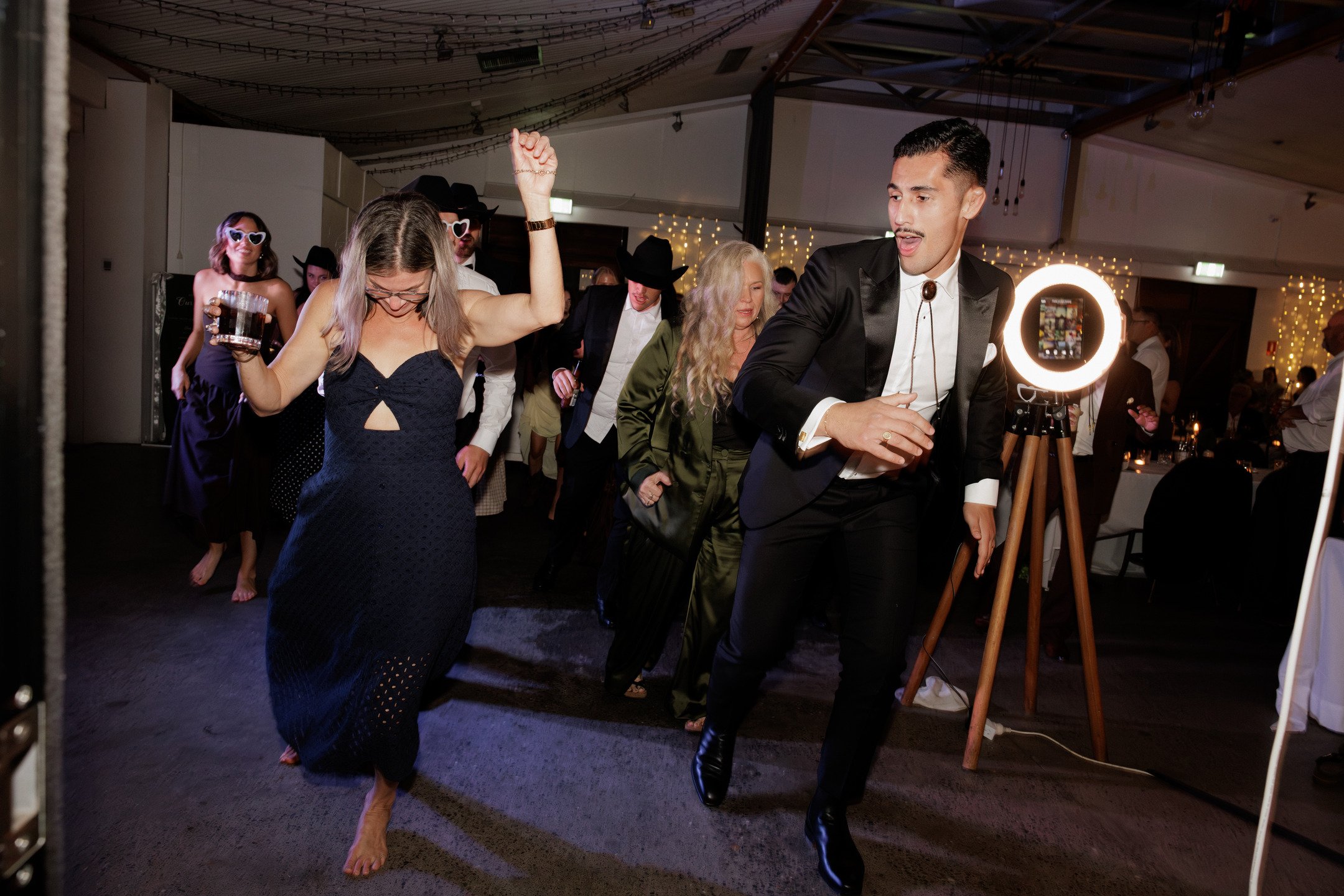 People dancing at a party, with some wearing sunglasses, formal attire, and enjoying the music and fun.