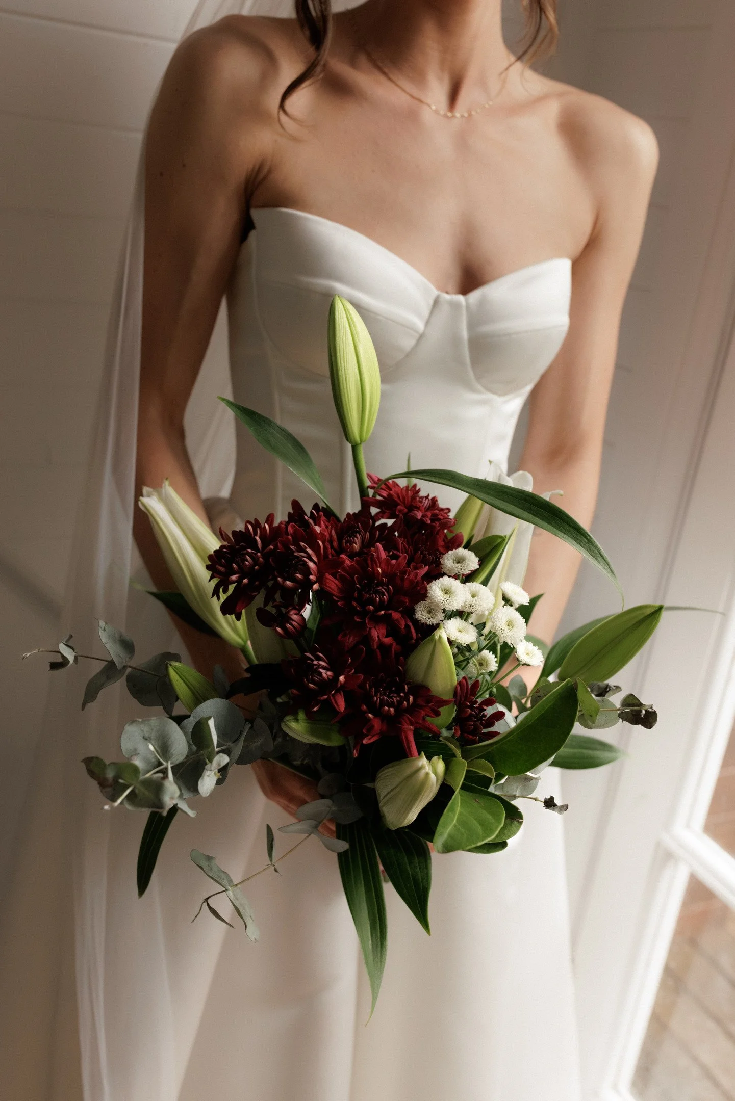 A woman in a white strapless dress holding a bouquet of flowers, including lilies, chrysanthemums, and greenery, standing indoors against a white wall.