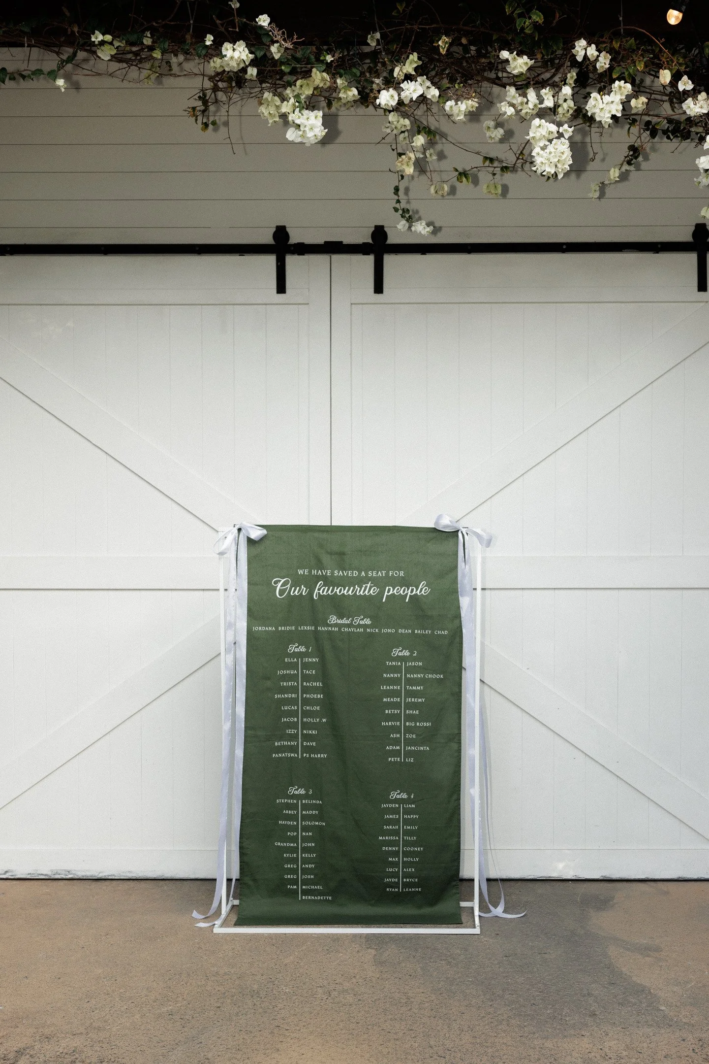 A seating chart for a wedding, displayed on a green fabric banner with white text and ribbons, hanging against a white wooden wall decorated with white flowers and foliage at the top.