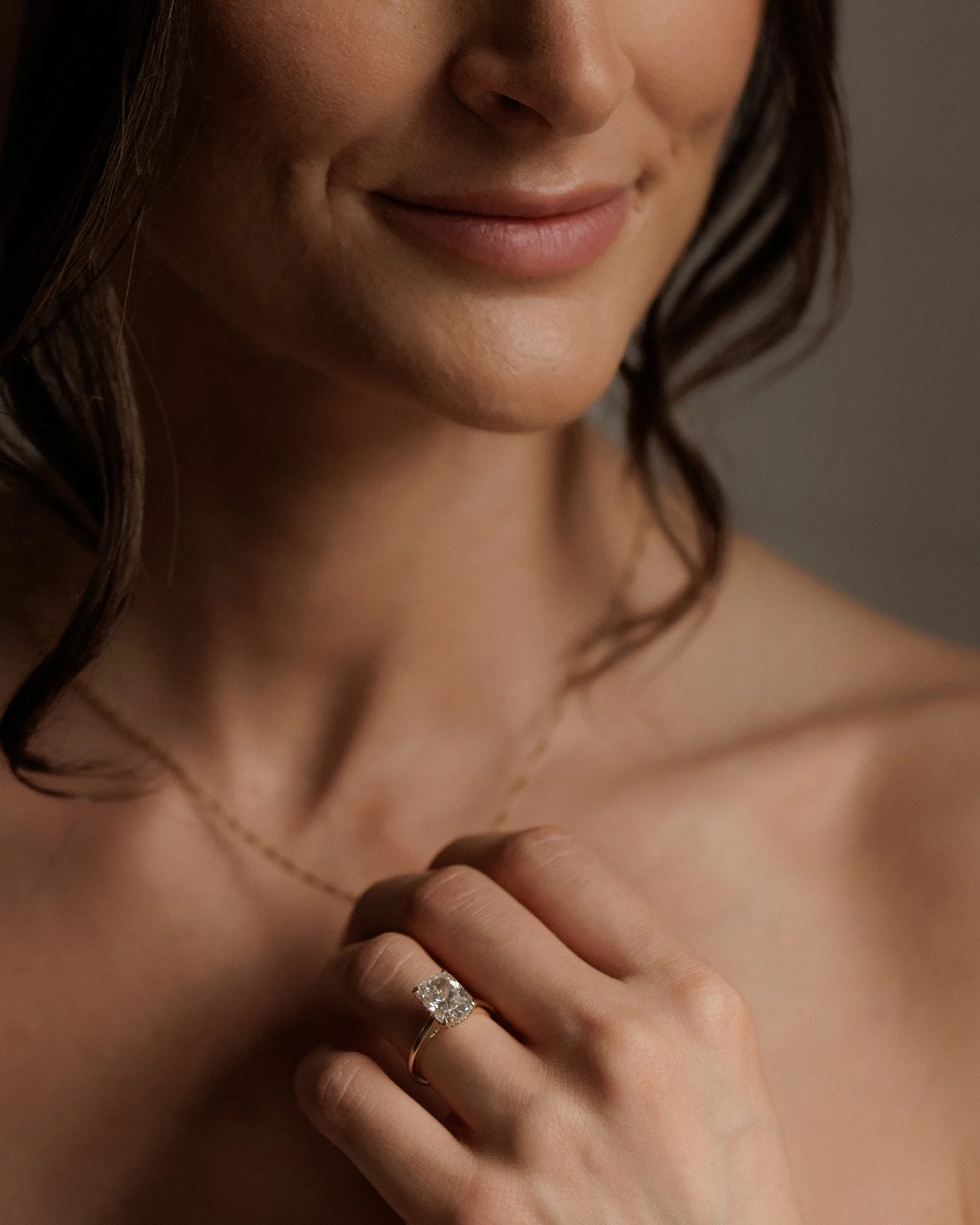 A close-up of a woman showing her face, neck, and hand. She is wearing a large diamond ring and a delicate necklace, with dark, wavy hair.