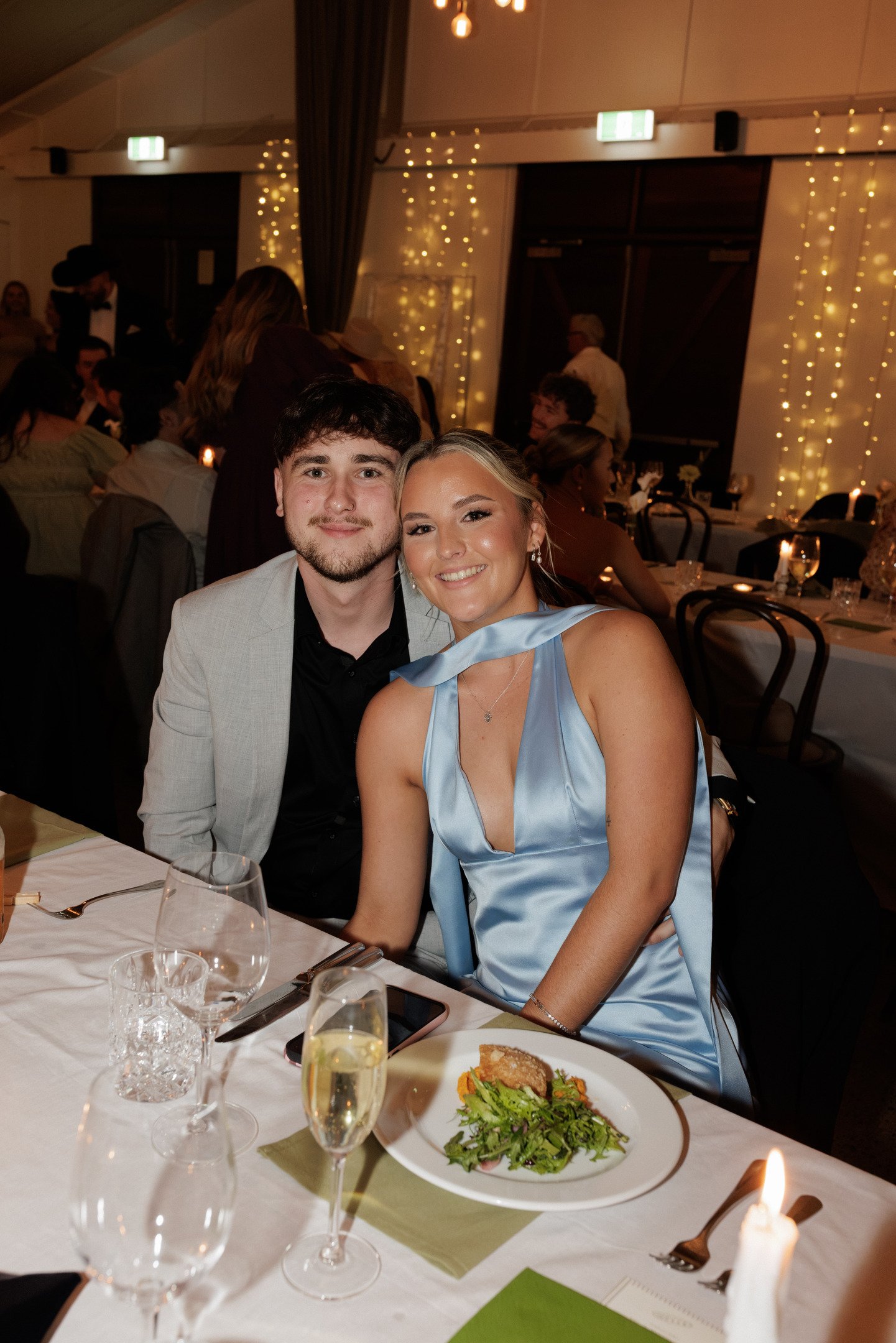 A young man and woman sitting at a table in a dimly lit banquet hall, smiling at the camera, with elegant tableware, a plate of salad, and a glass of champagne in front of them. The background features other guests, fairy lights, and a festive atmosp