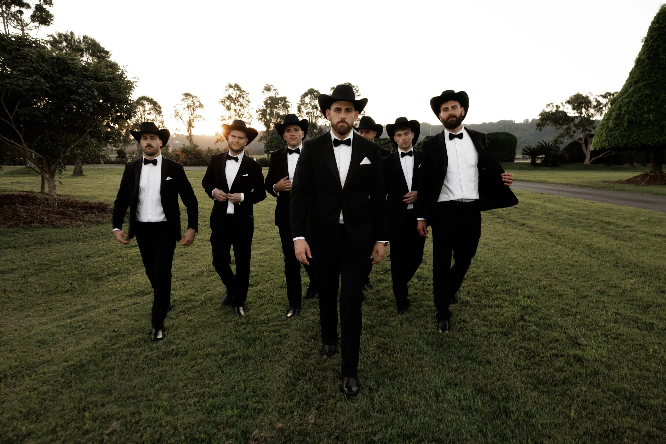 A group of six men dressed in black tuxedos with bow ties and white shirts, wearing black cowboy hats, walking together outdoors in a park at sunset.