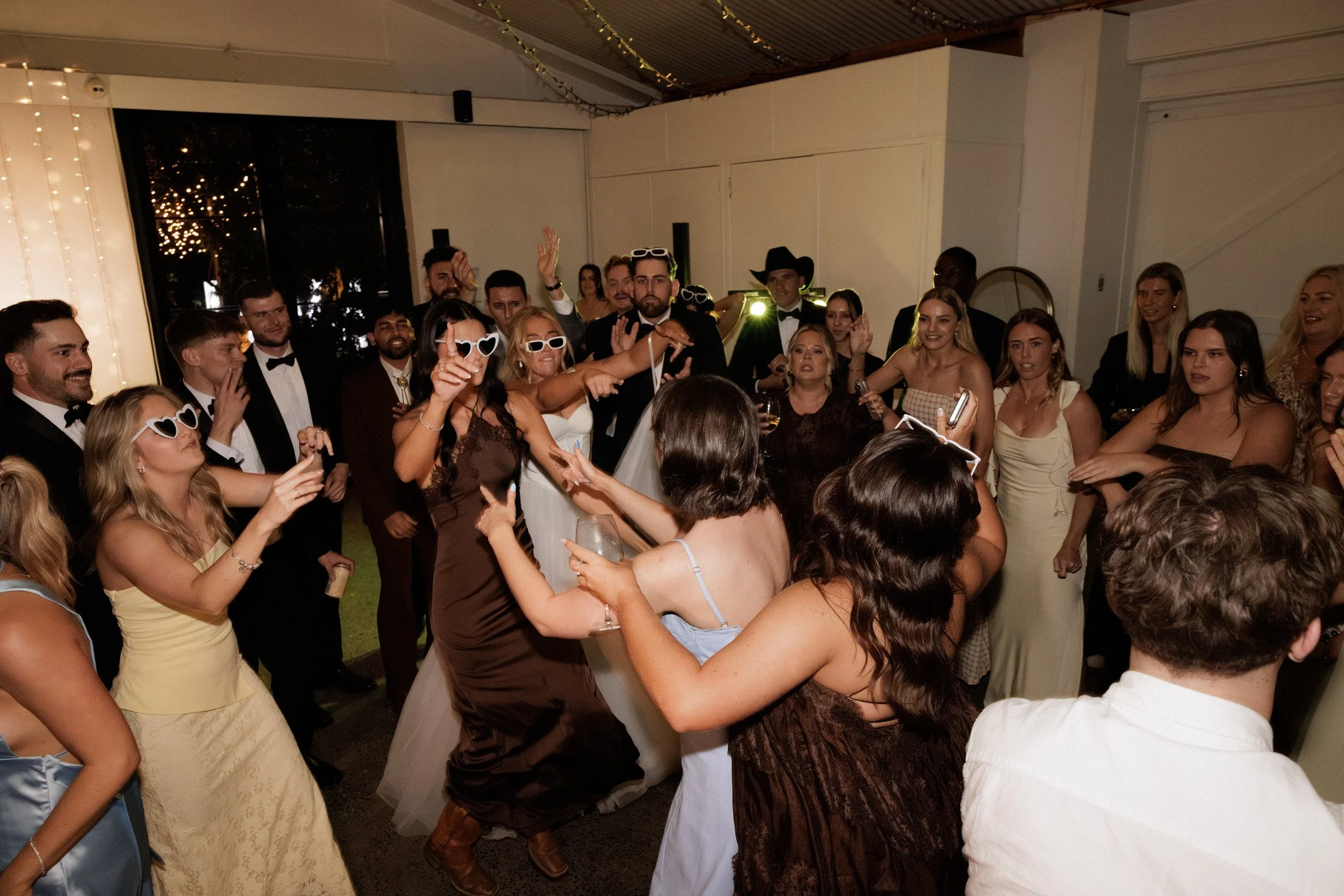 A group of people dancing and celebrating at an indoor party or wedding reception. Many are dressed in formal attire, with some wearing sunglasses and others in tuxedos or dresses. The scene is lively and festive.