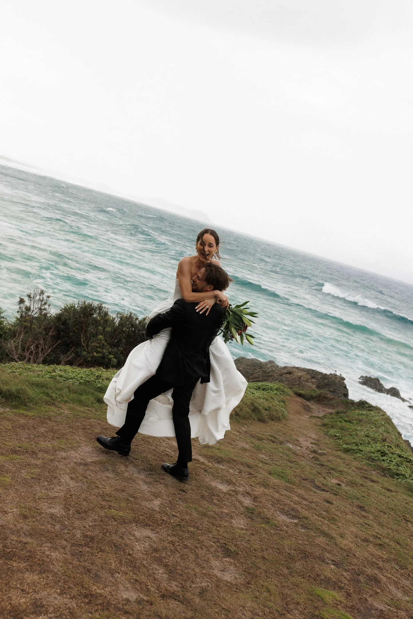 A man in a black suit lifting a woman in a white dress at a beachside location, with wave-covered shoreline in the background and cloudy sky.