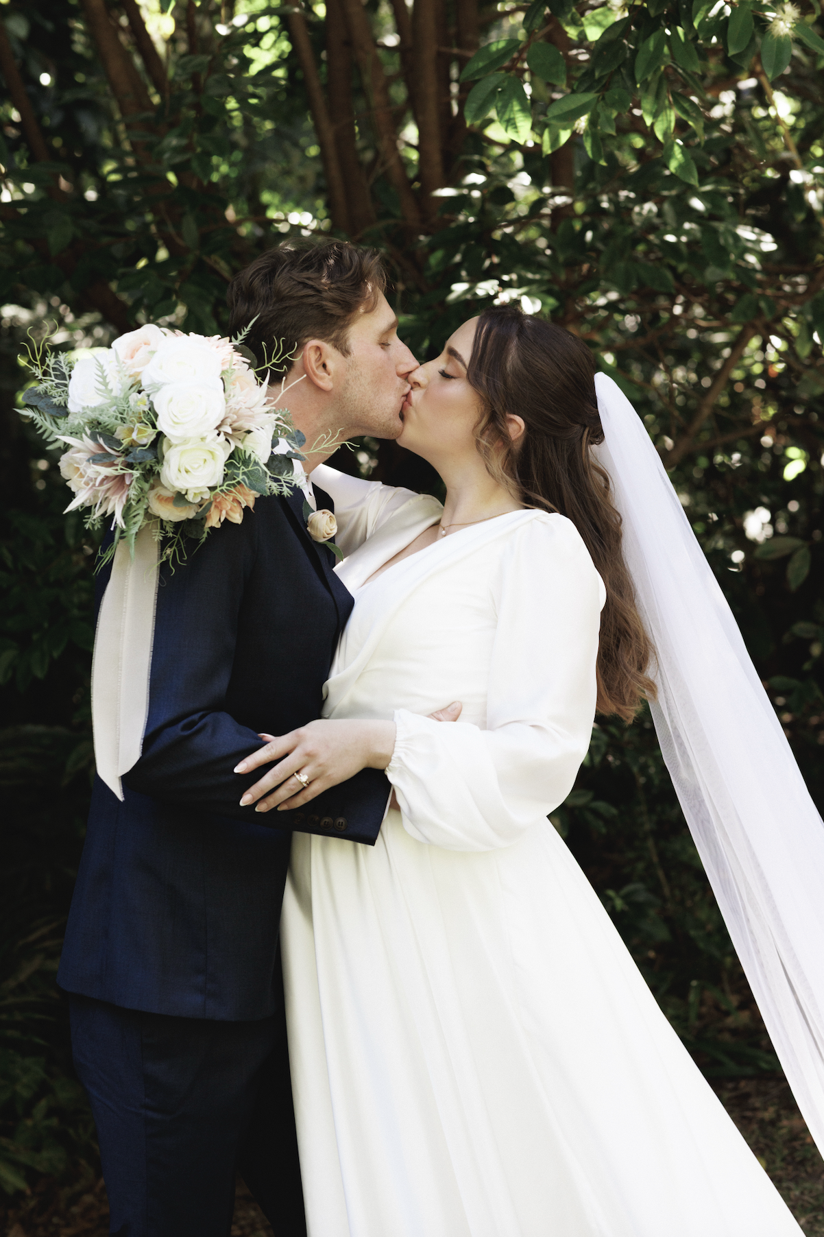 A newlywed couple is kissing outdoors, surrounded by green foliage. The groom is holding a bouquet of white and blush flowers, and the bride is wearing a white dress with a veil.
