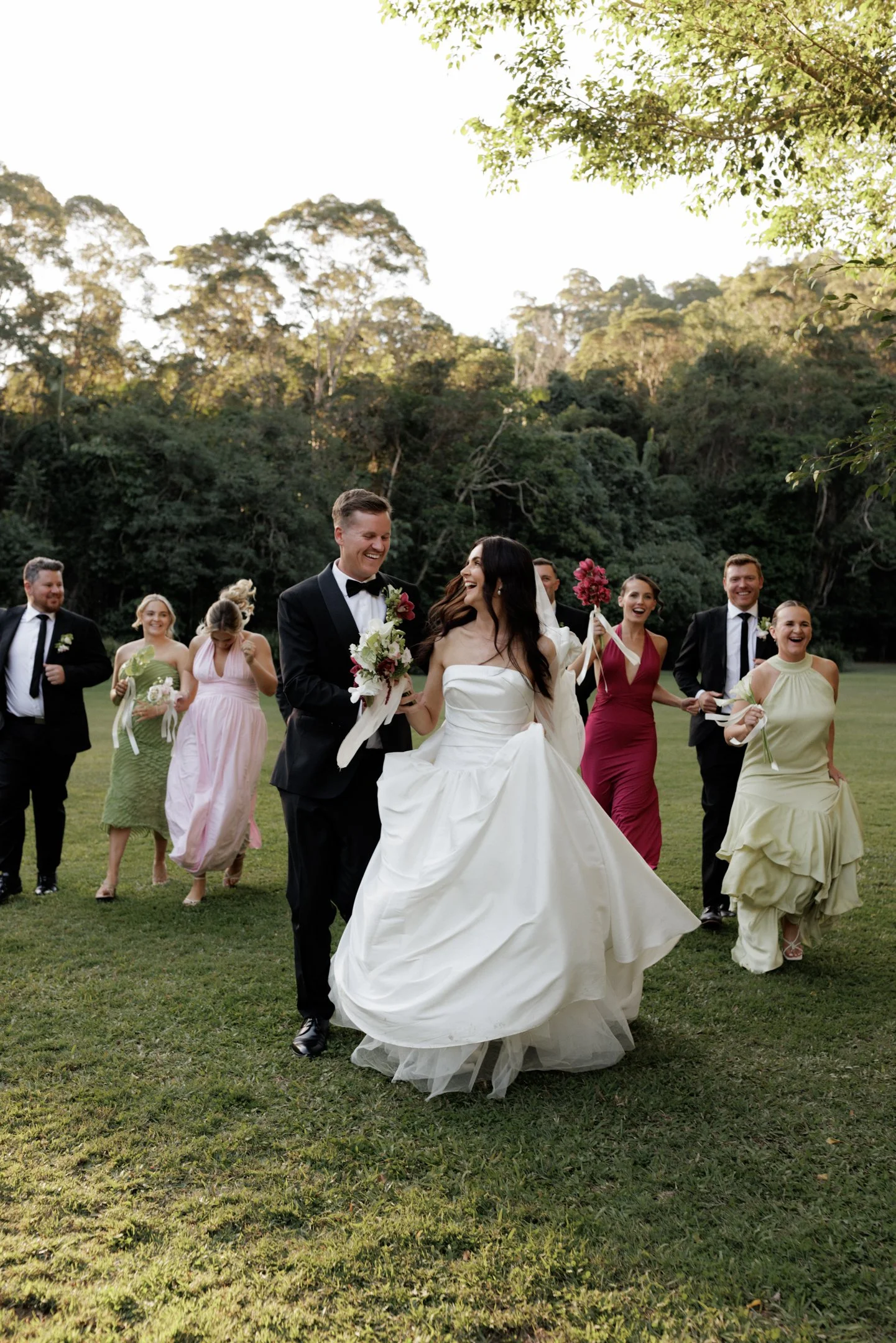 A group of people, including a bride in a white wedding gown and groom in a black tuxedo, are walking outdoors on grass, celebrating a wedding. The group is smiling and joyful, with some holding flower bouquets. The background features lush green tre