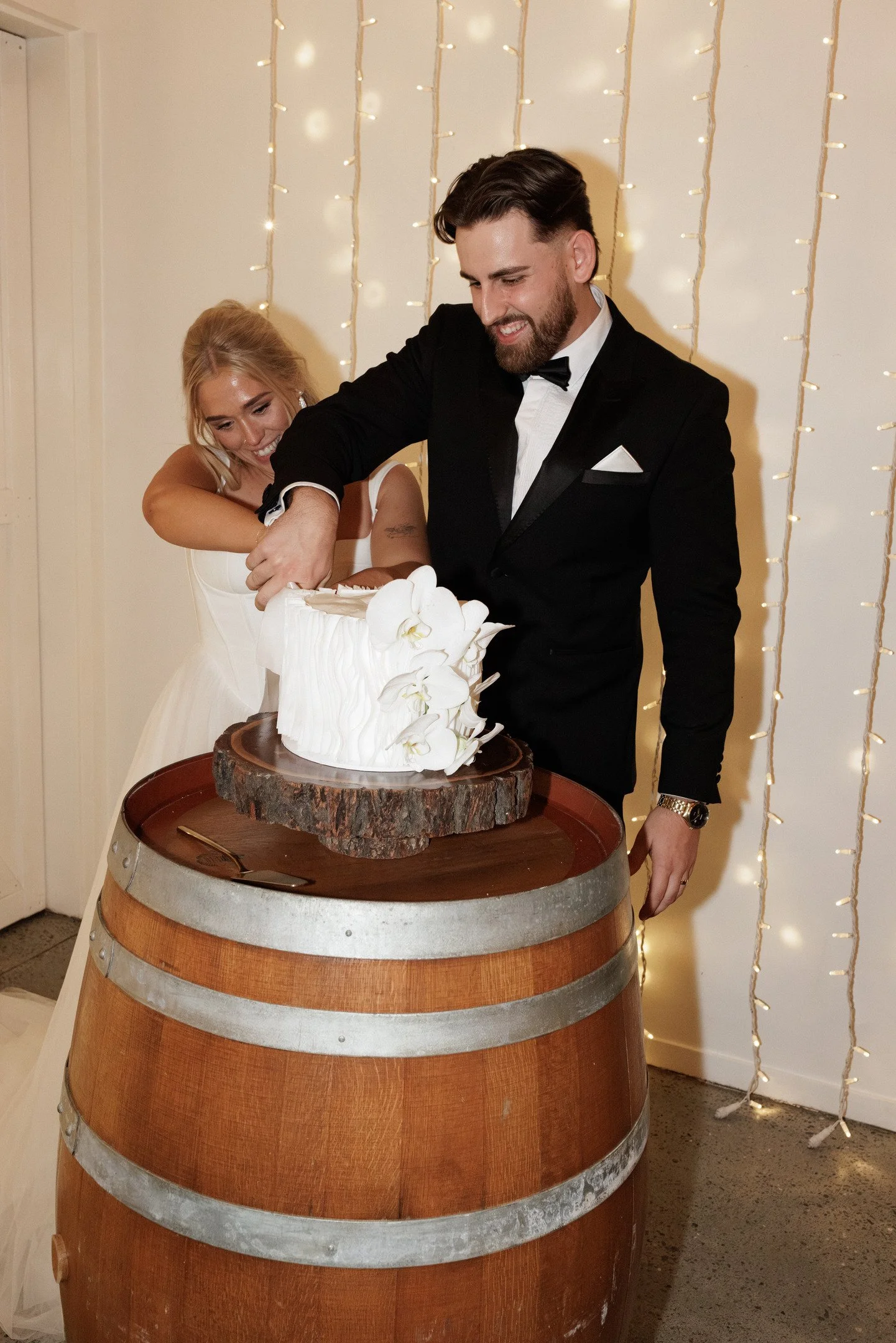 A couple in formal wedding attire cutting a wedding cake together, with decorative string lights in the background, located on a wine barrel serving as a table.