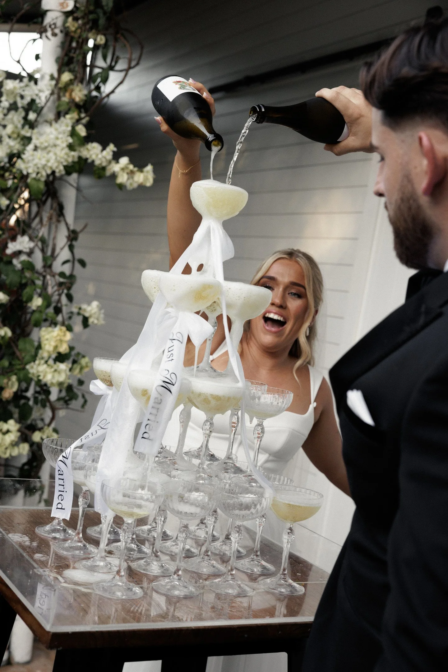 A wedding reception scene with a bottle pouring champagne into a glass held by a woman, with a multi-tiered champagne tower and a man in a tuxedo nearby, outdoors with floral decorations.