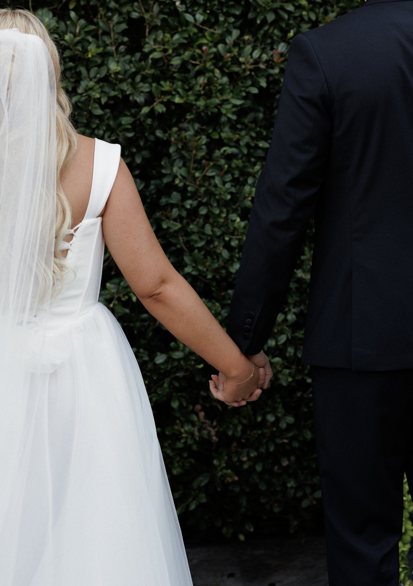 A bride and groom holding hands during their wedding ceremony outdoors, with a background of green bushes.