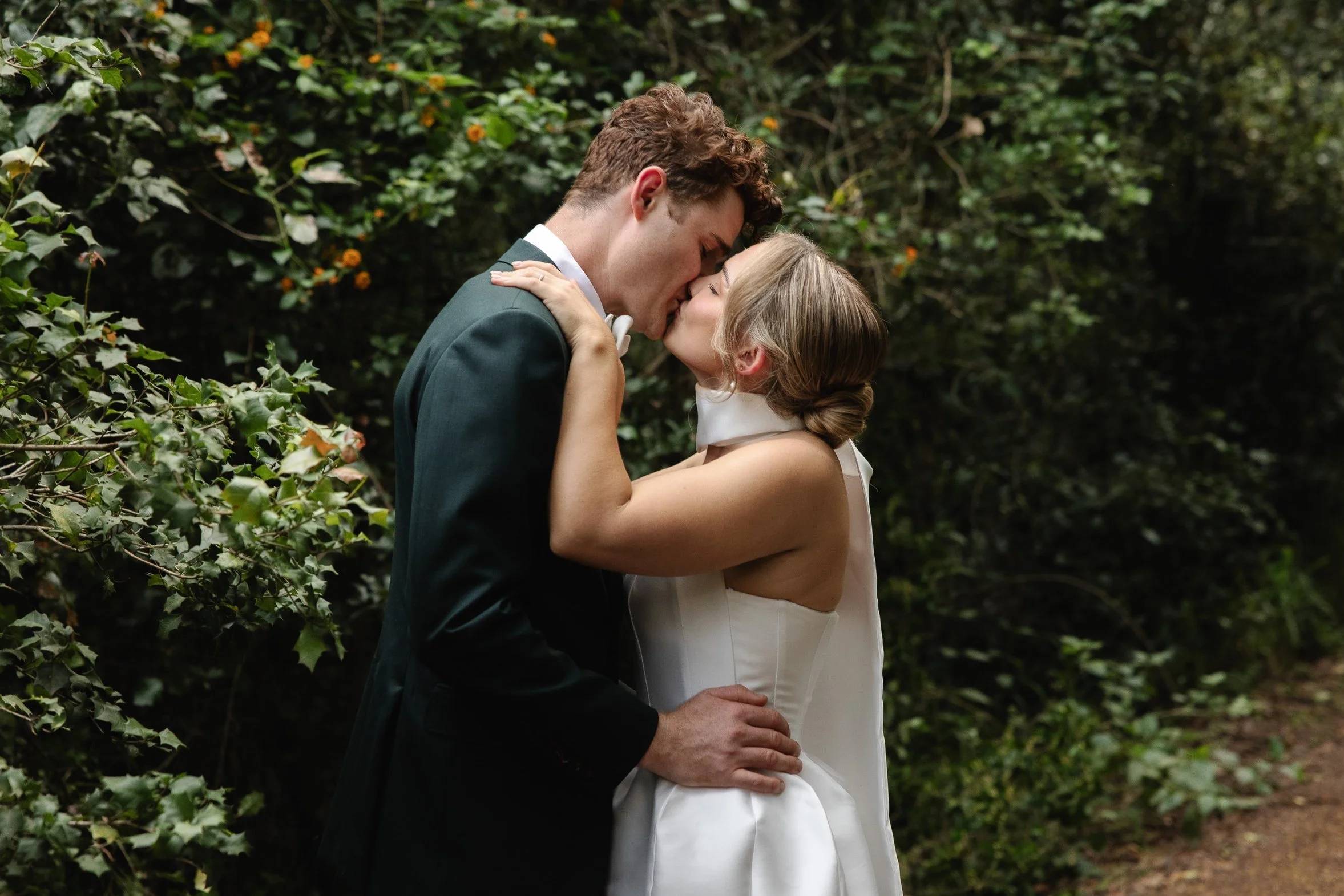 A bride and groom sharing a kiss outdoors among green foliage.