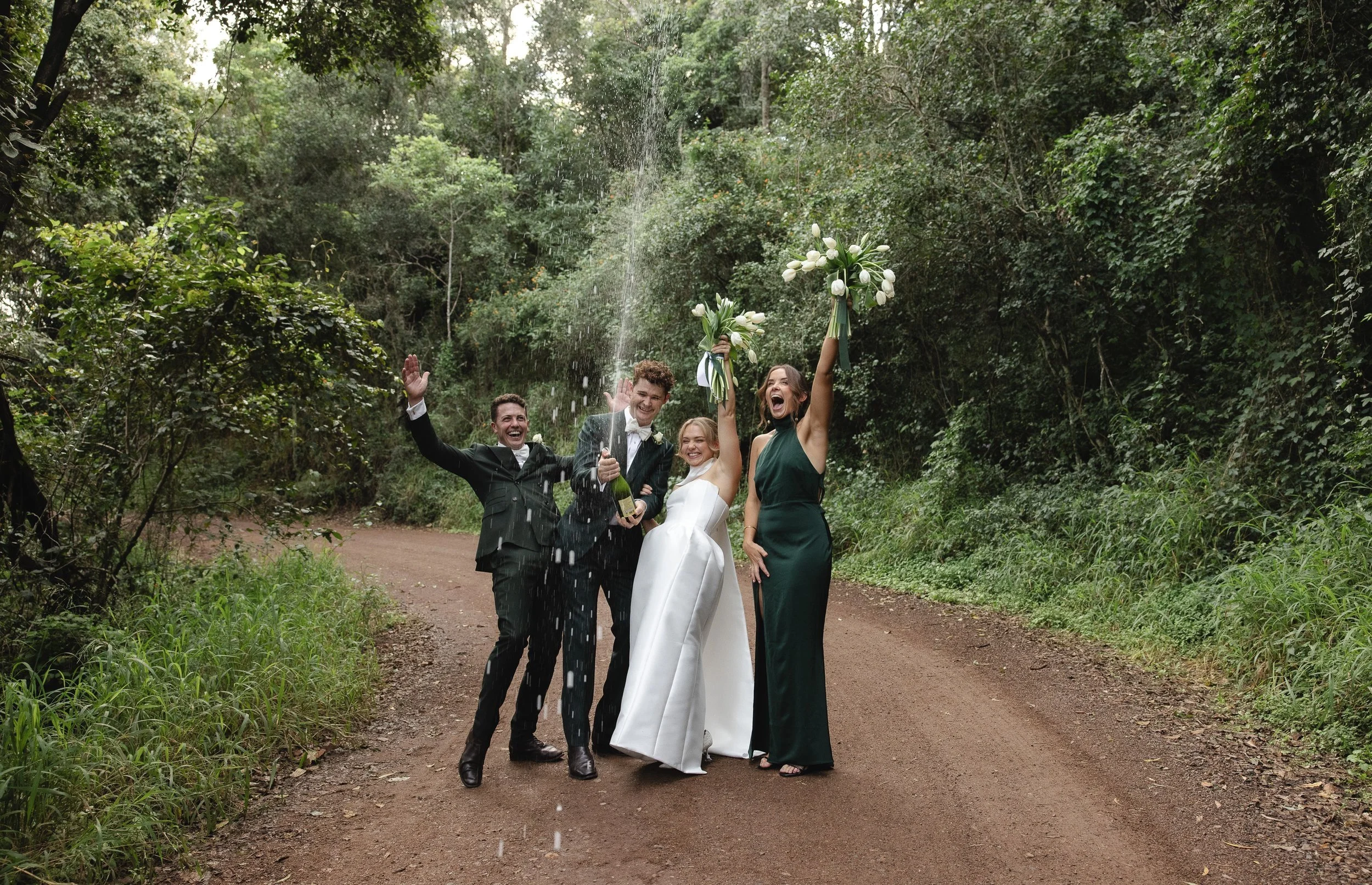 Group of four people celebrating on a forest dirt path, with the bride holding a bouquet and the others raising their arms, one person spraying champagne, surrounded by lush green trees.
