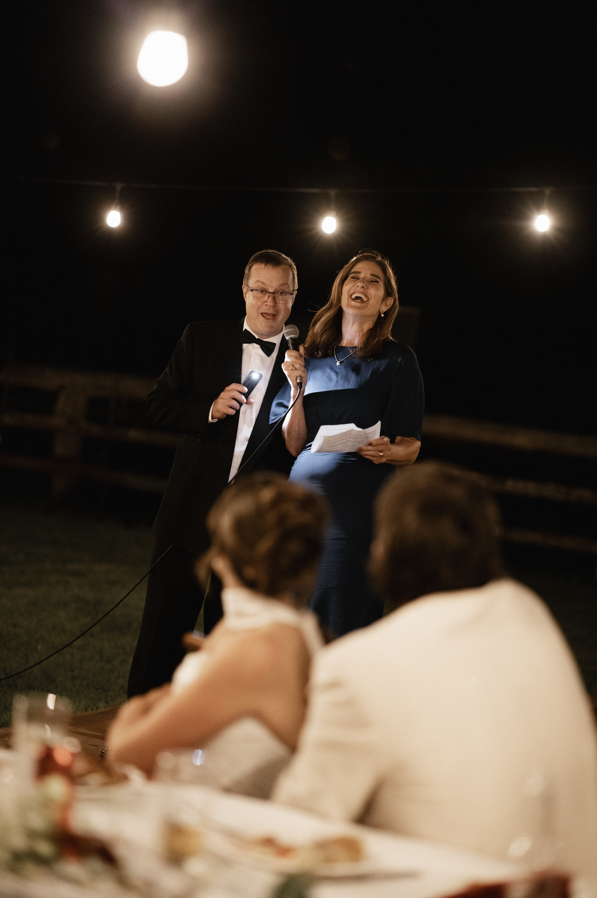 A man in a tuxedo and a woman in a dark dress are singing and holding papers at an outdoor nighttime event, with string lights overhead and people seated at a table in front.