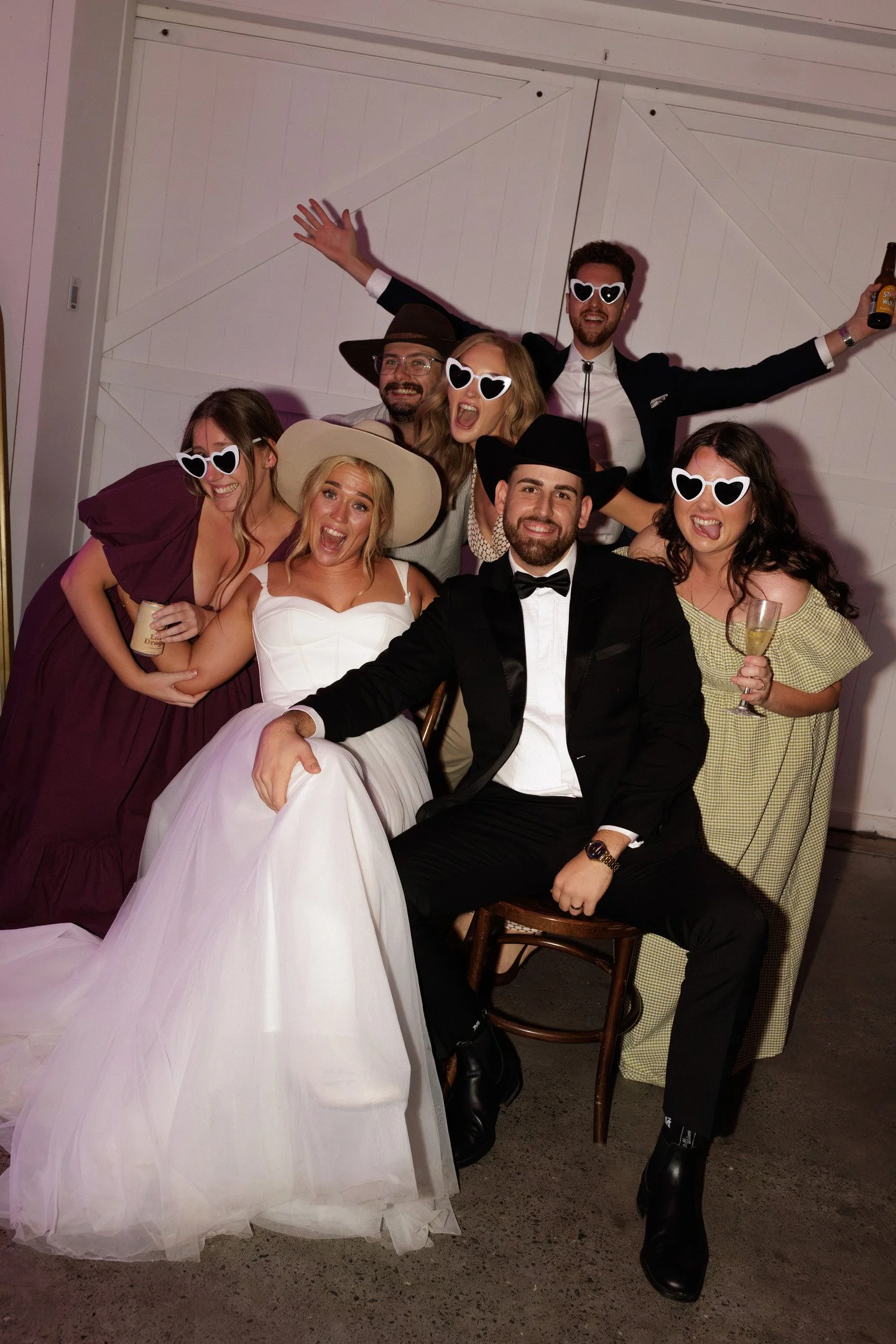 Group of people at a celebration or party, wearing sunglasses, some in formal attire, all smiling and having fun. One person is holding a beer, and another holding a glass of champagne. The background appears to be a white barn or garage door.