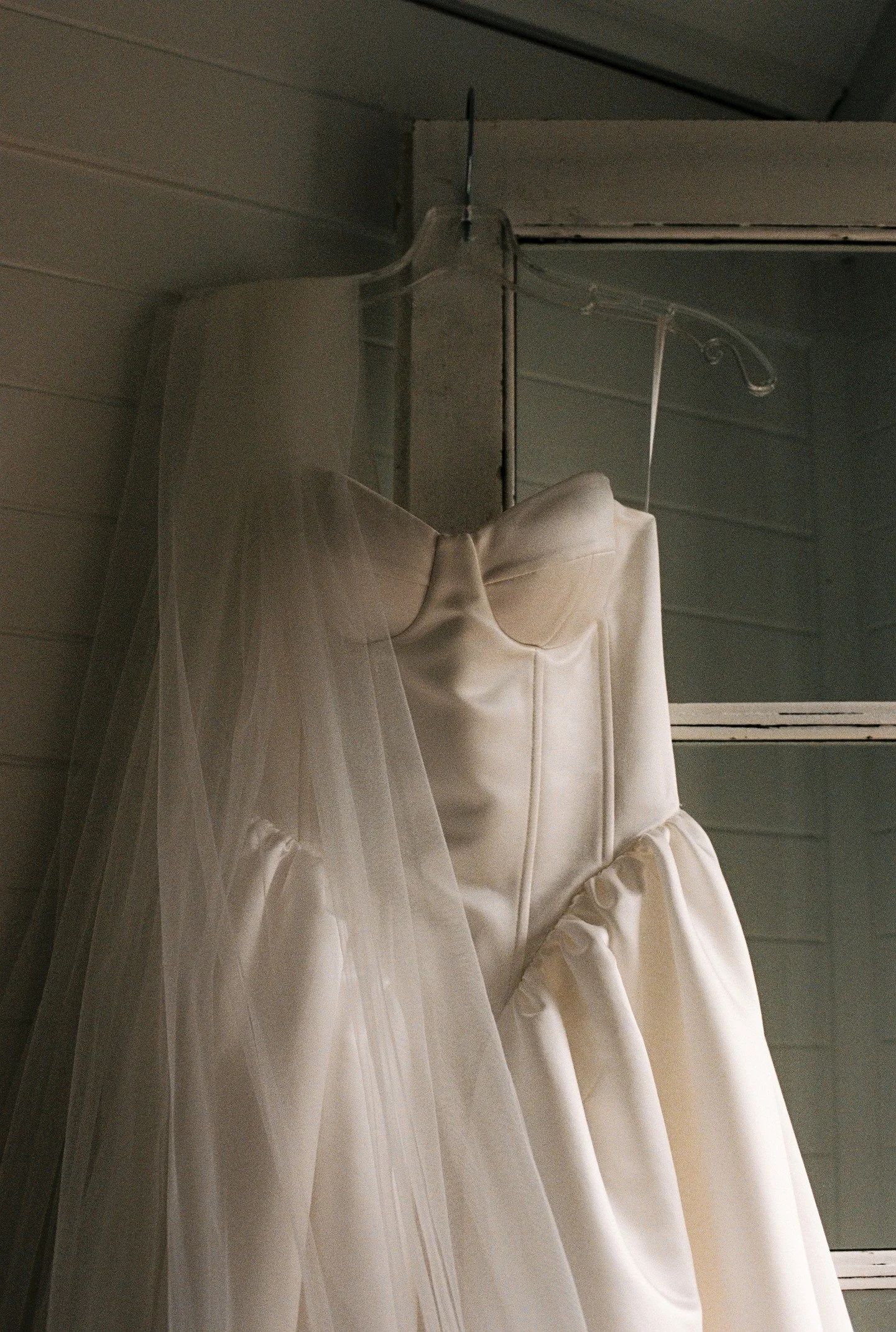 A white wedding dress with a veil hanging on a hanger, placed against a wall with horizontal wooden paneling.
