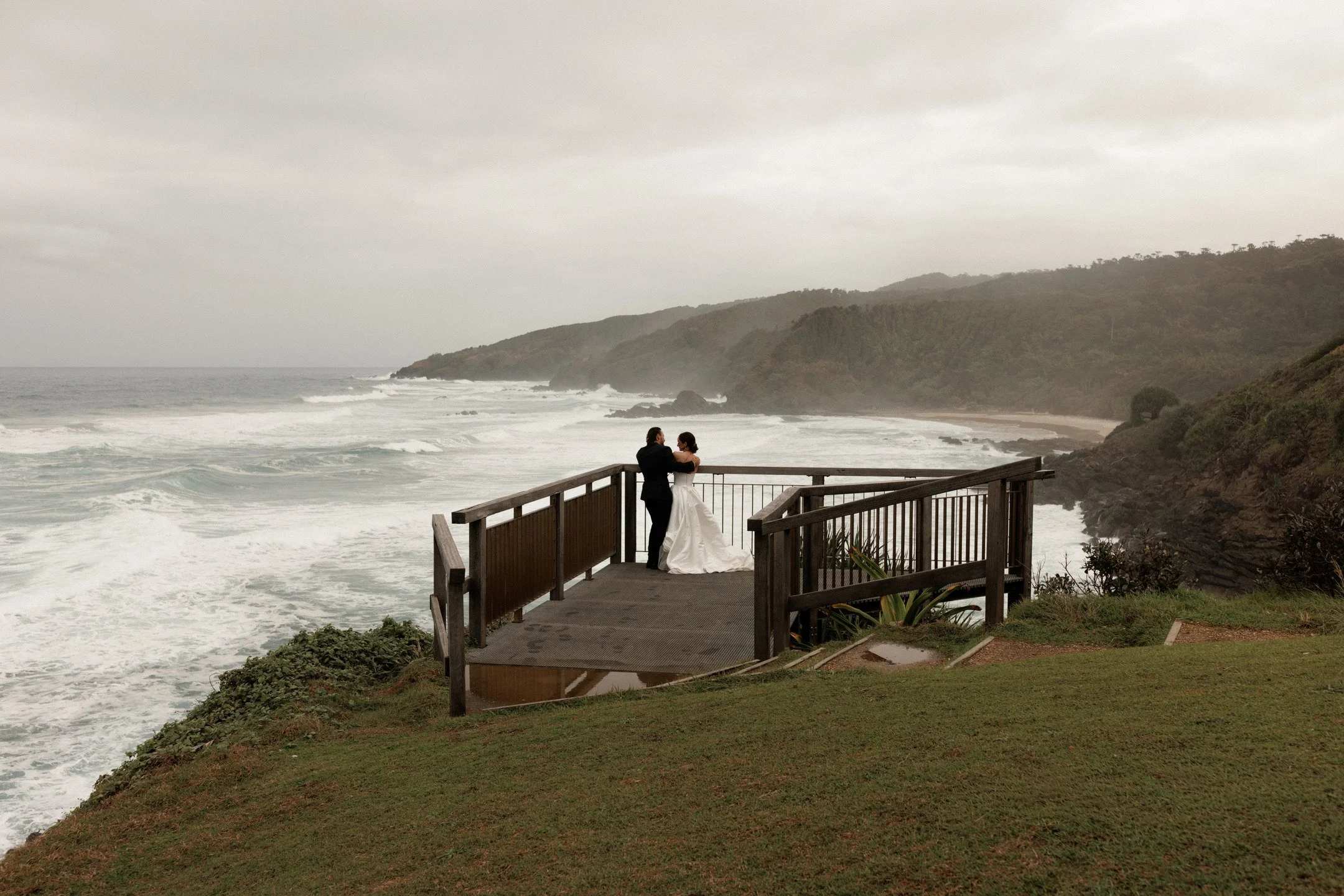 A couple dressed in wedding attire standing on a wooden observation deck overlooking the ocean, with waves crashing on the shore and green hills in the background under a cloudy sky.
