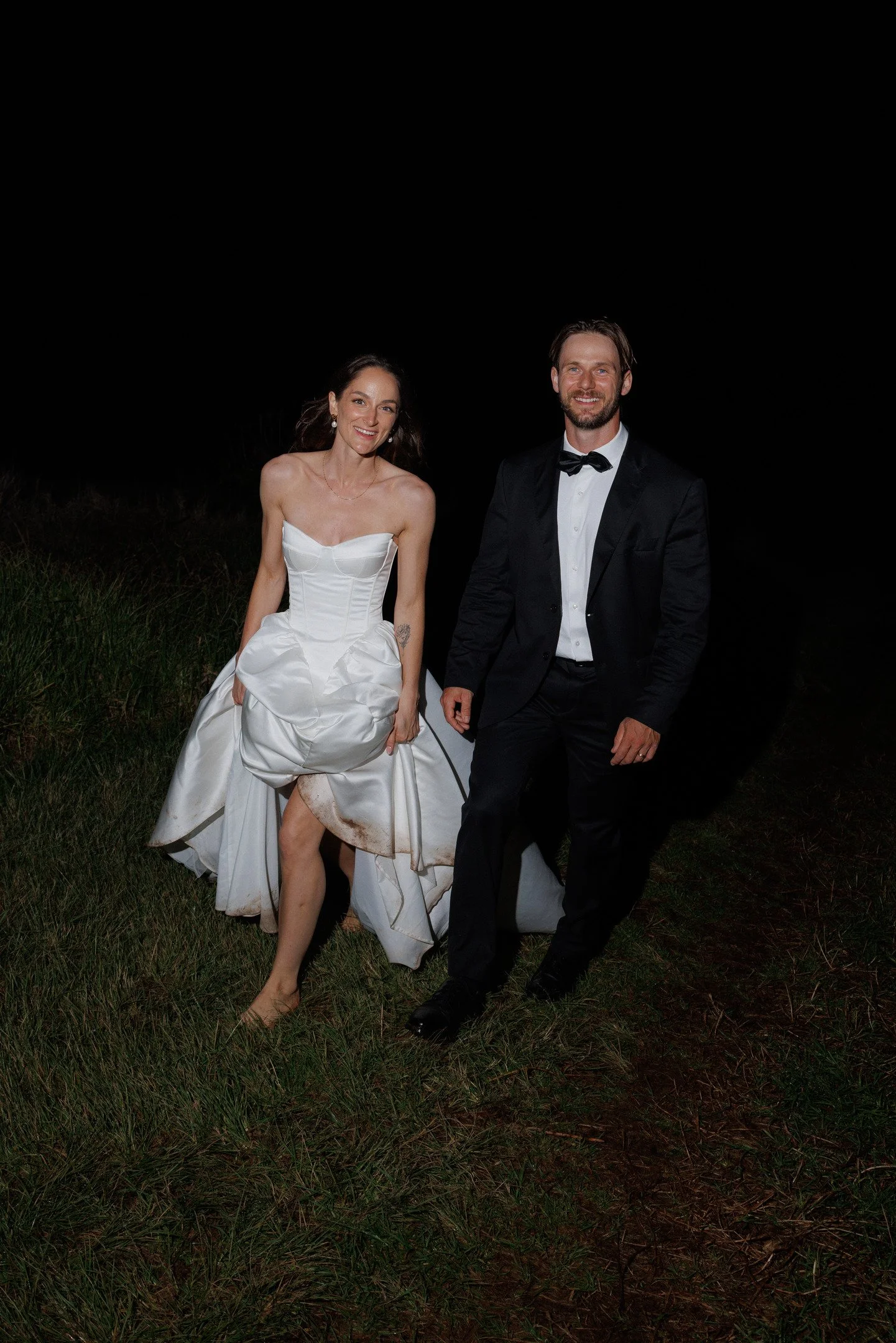 A happy woman in a wedding dress and a happy man in a tuxedo are standing outdoors at night. The woman is barefoot, holding her dress up slightly, showing her legs and a tattoo on her arm. The man is smiling, standing next to her with his hands at hi