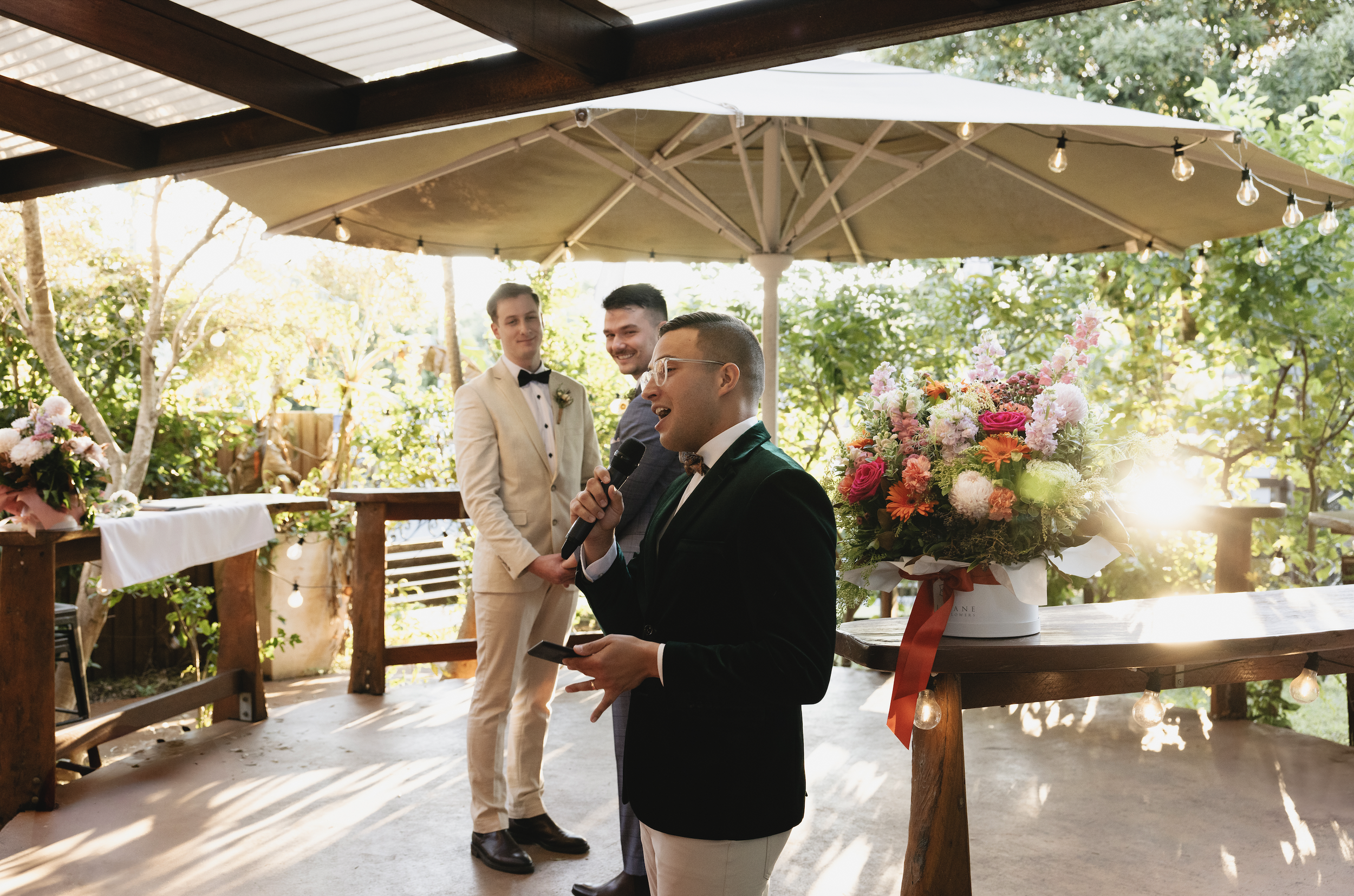 A person in a black tuxedo gives a speech at a wedding reception outdoors on a patio with wooden tables, flowers, and string lights, with two men in suits standing behind him.