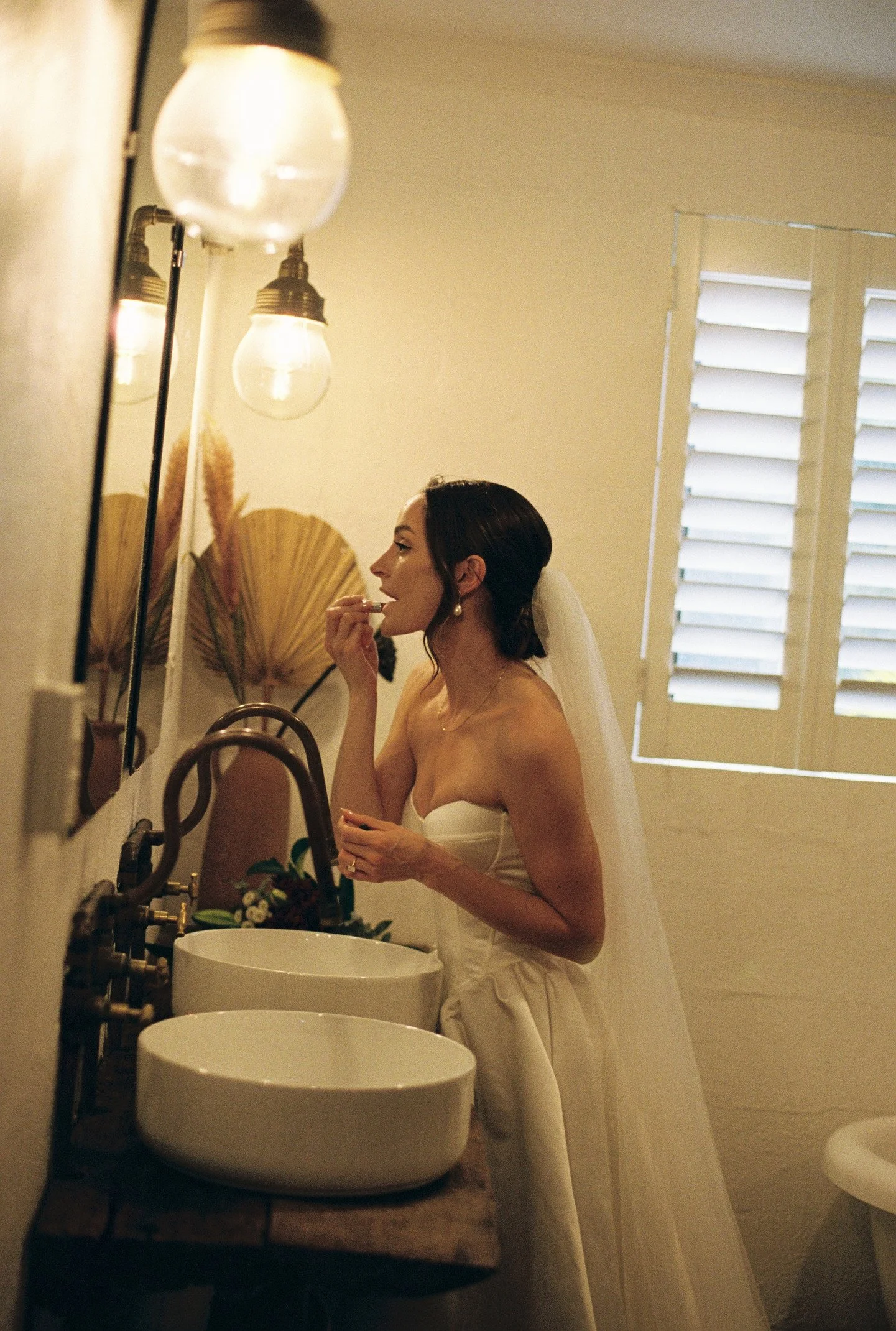 A woman in a wedding dress applying lipstick in front of a mirror in a bathroom with natural light coming through the window.